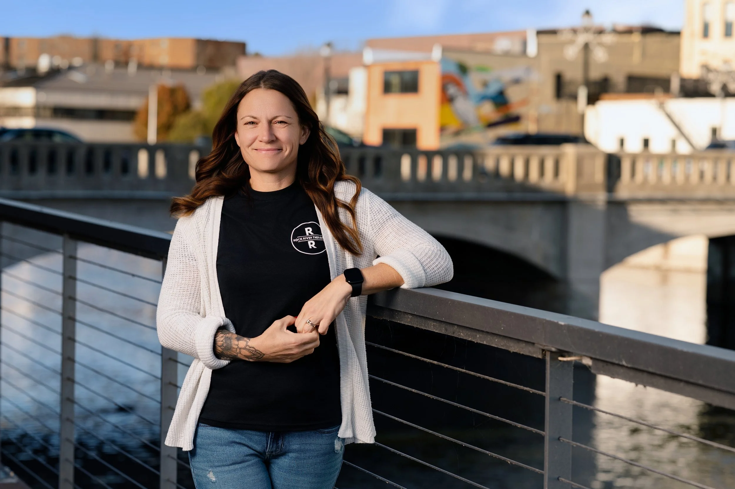Woman with brown hair, wearing a black shirt, white cardigan, and blue jeans, leaning on a railing by a river with a bridge in the background during daytime.