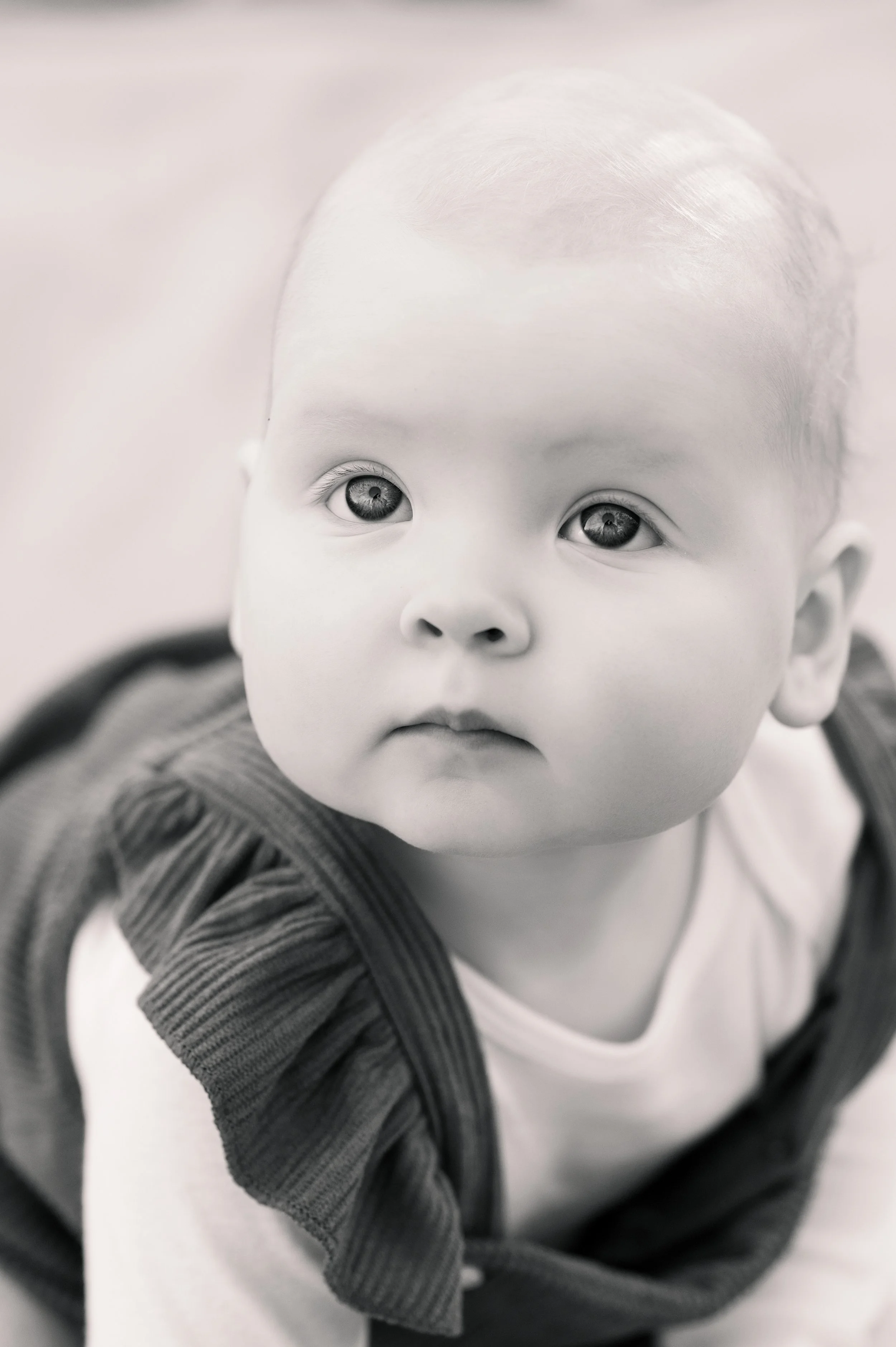 A black-and-white close-up photograph of a baby with big, expressive eyes, wearing a light-colored shirt and a dark, textured jacket.