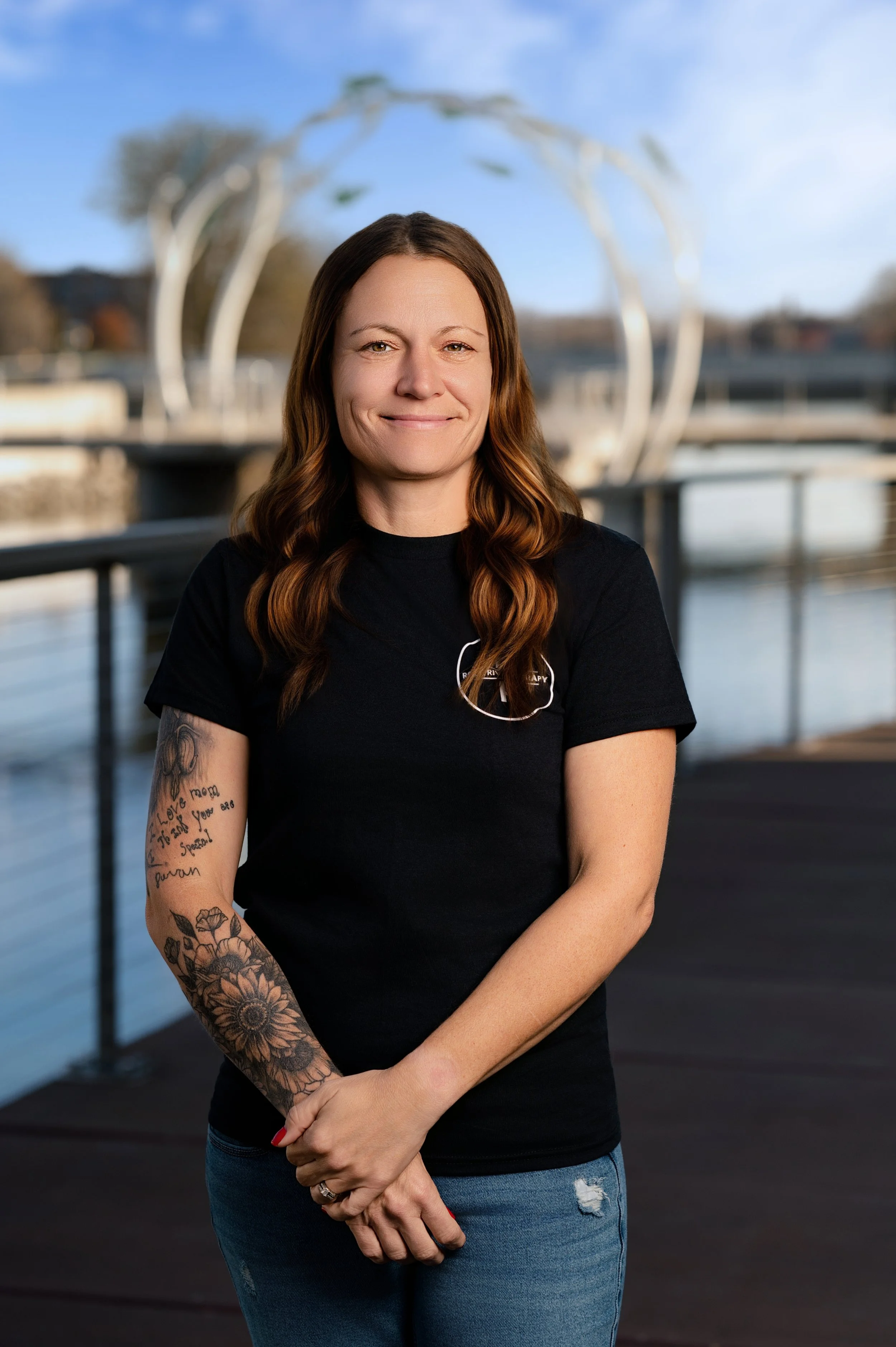 A woman with long wavy brown hair, wearing a black T-shirt and blue jeans, standing on a dock near water with a modern sculpture in the background.