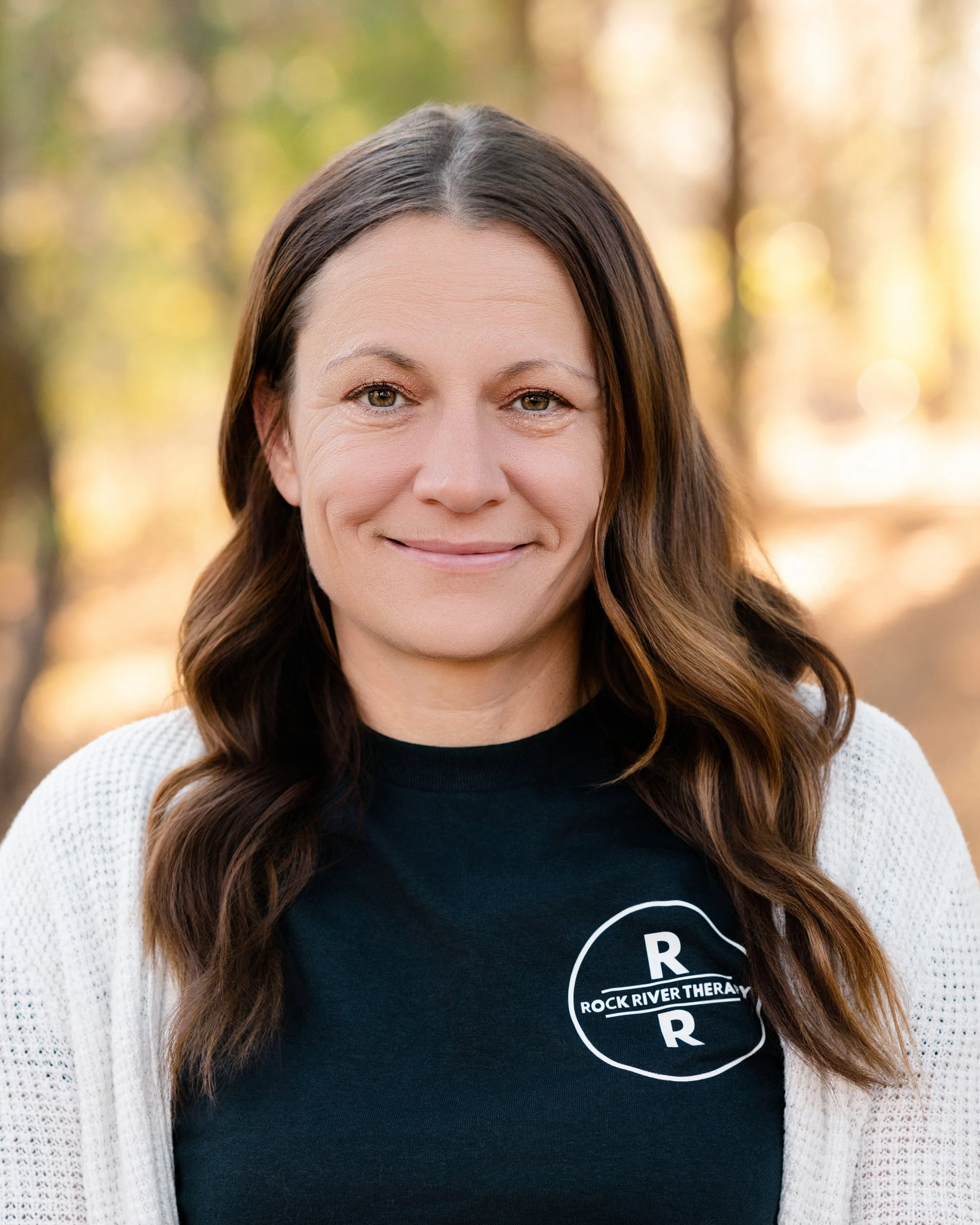A woman with long brown hair smiling outdoors during autumn, wearing a black shirt with a logo that reads 'Rock River Therapy' and a white cardigan.