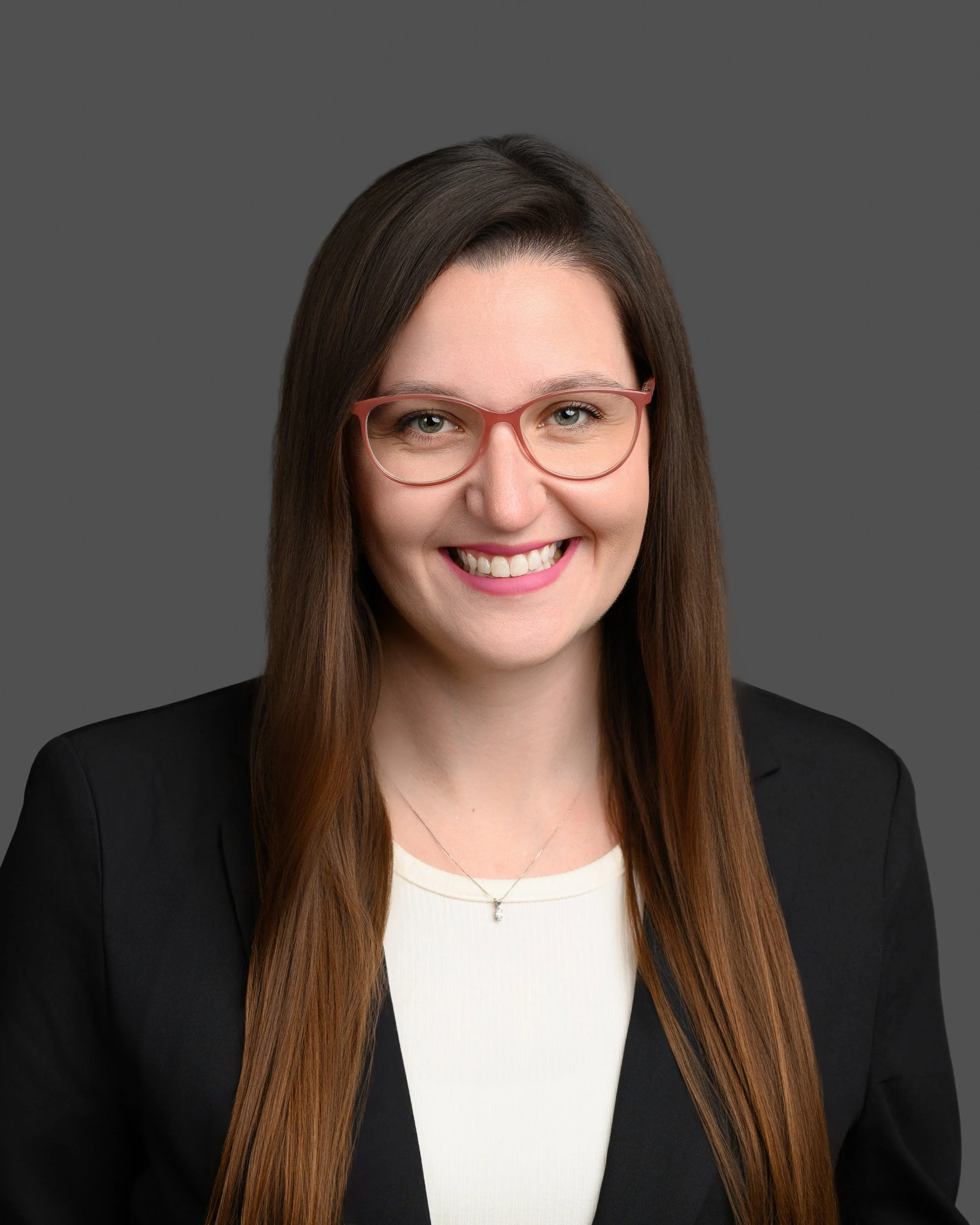 A smiling woman with long brown hair, wearing pink glasses, a black blazer, and a white top, against a gray background.