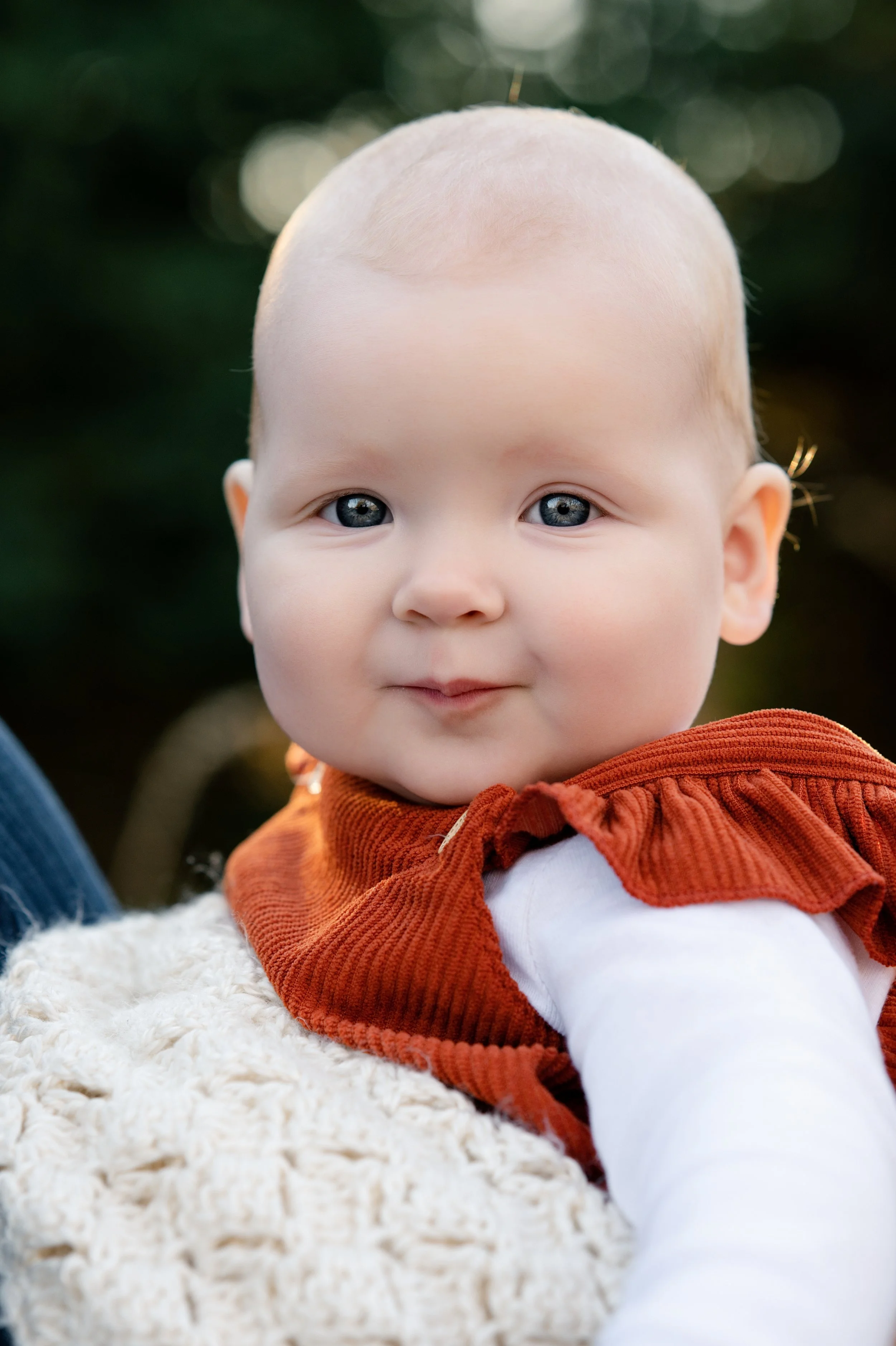 Close-up of a baby with blue eyes, wearing an orange vest and white long-sleeve shirt, outdoors with a blurred green background.