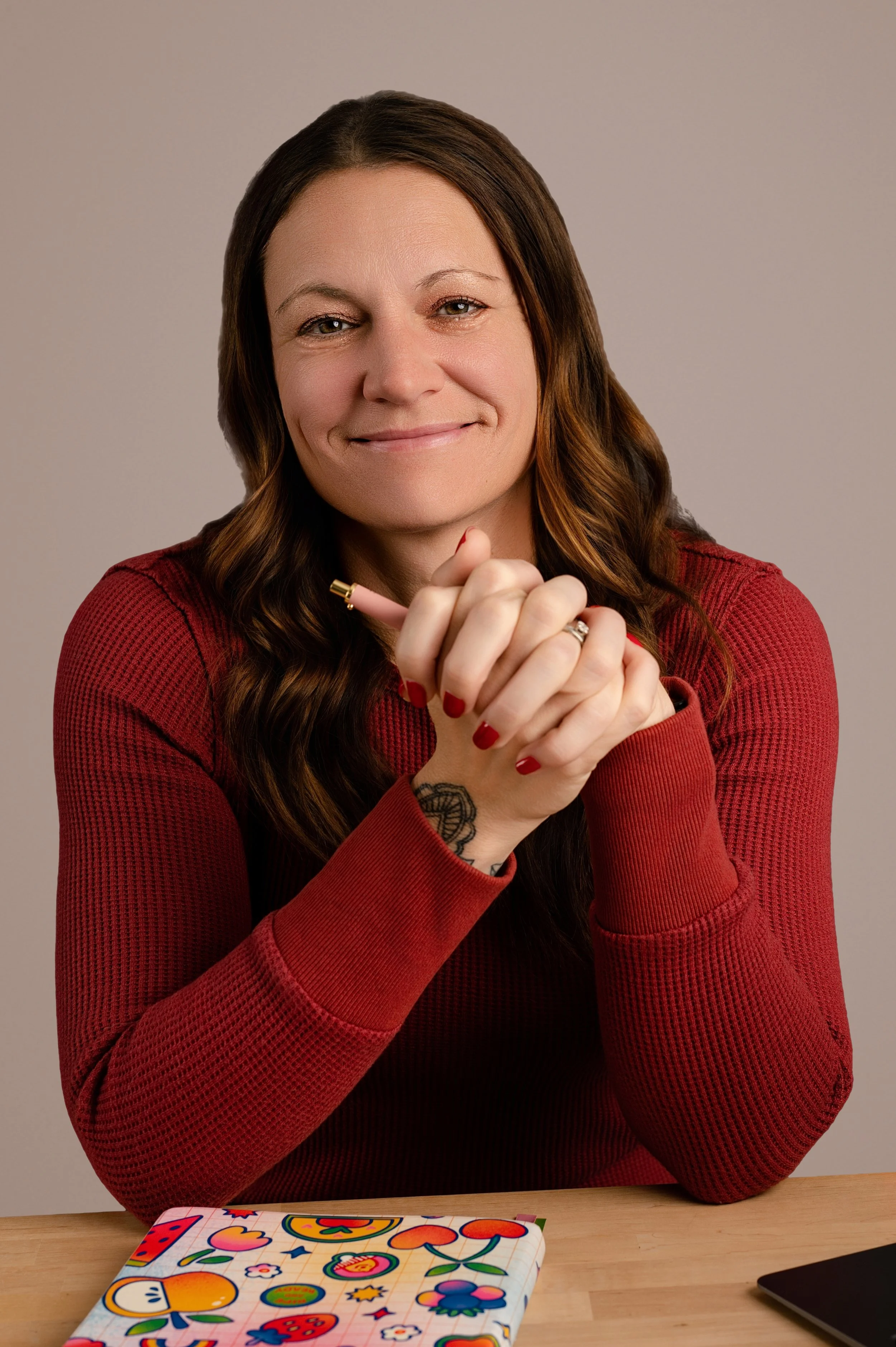 A woman with long brown hair and light skin wearing a red sweater, sitting at a desk with colorful stickers or drawings on a notepad in front of her. She is smiling, with her hands clasped together in front of her, holding a pink pencil.