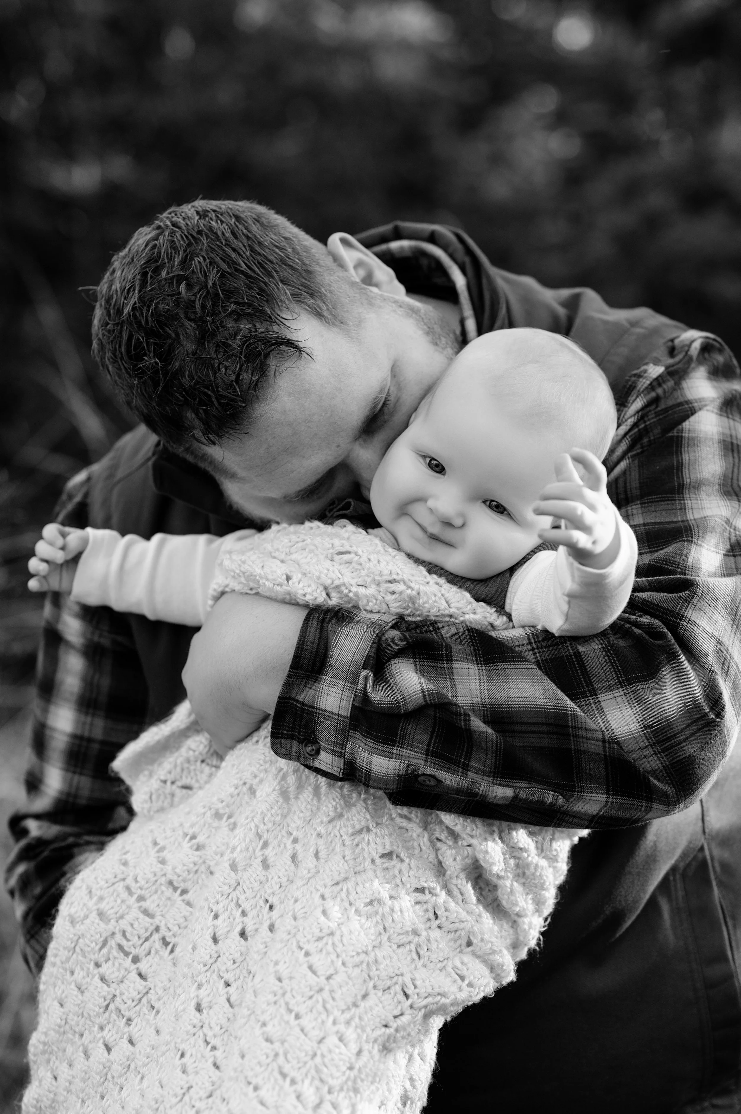 A man cuddles a baby with a gentle kiss on the head outdoors, both wrapped in a cozy blanket, in black and white.