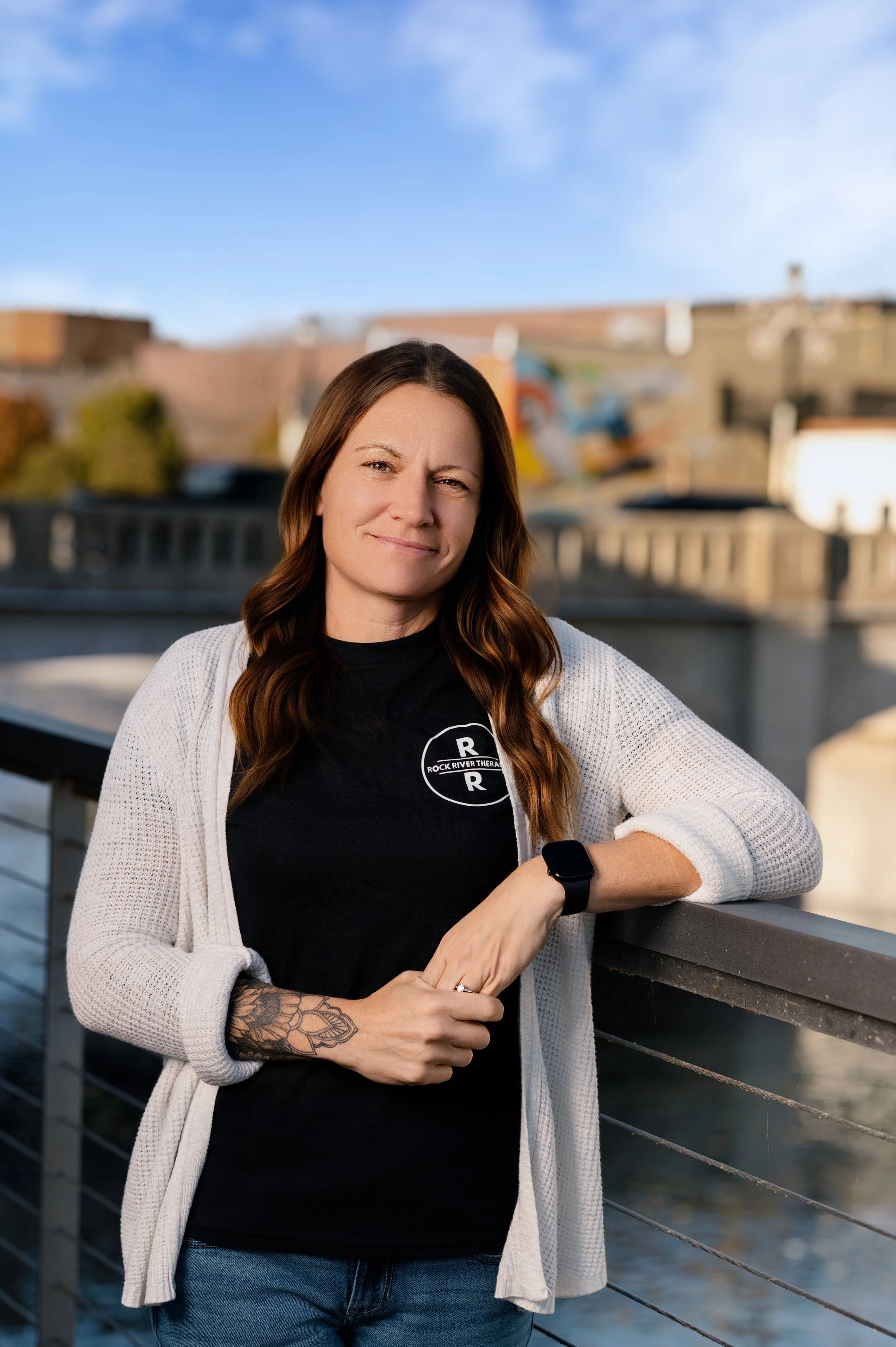 A woman with wavy brown hair standing on a bridge outdoors, wearing a black t-shirt with 'Rock River Therapy' logo, a white cardigan, and a smartwatch, with a river and cityscape in the background.