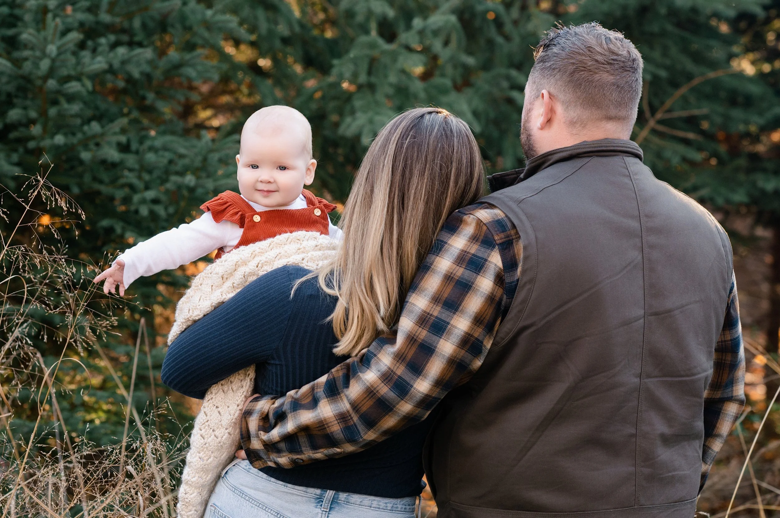 A family outdoors in a wooded area during autumn, with a young child being held by a woman and a man standing nearby. The child is looking at the camera and smiling, wearing a rust-colored dress with ruffle straps over a white long-sleeve shirt.