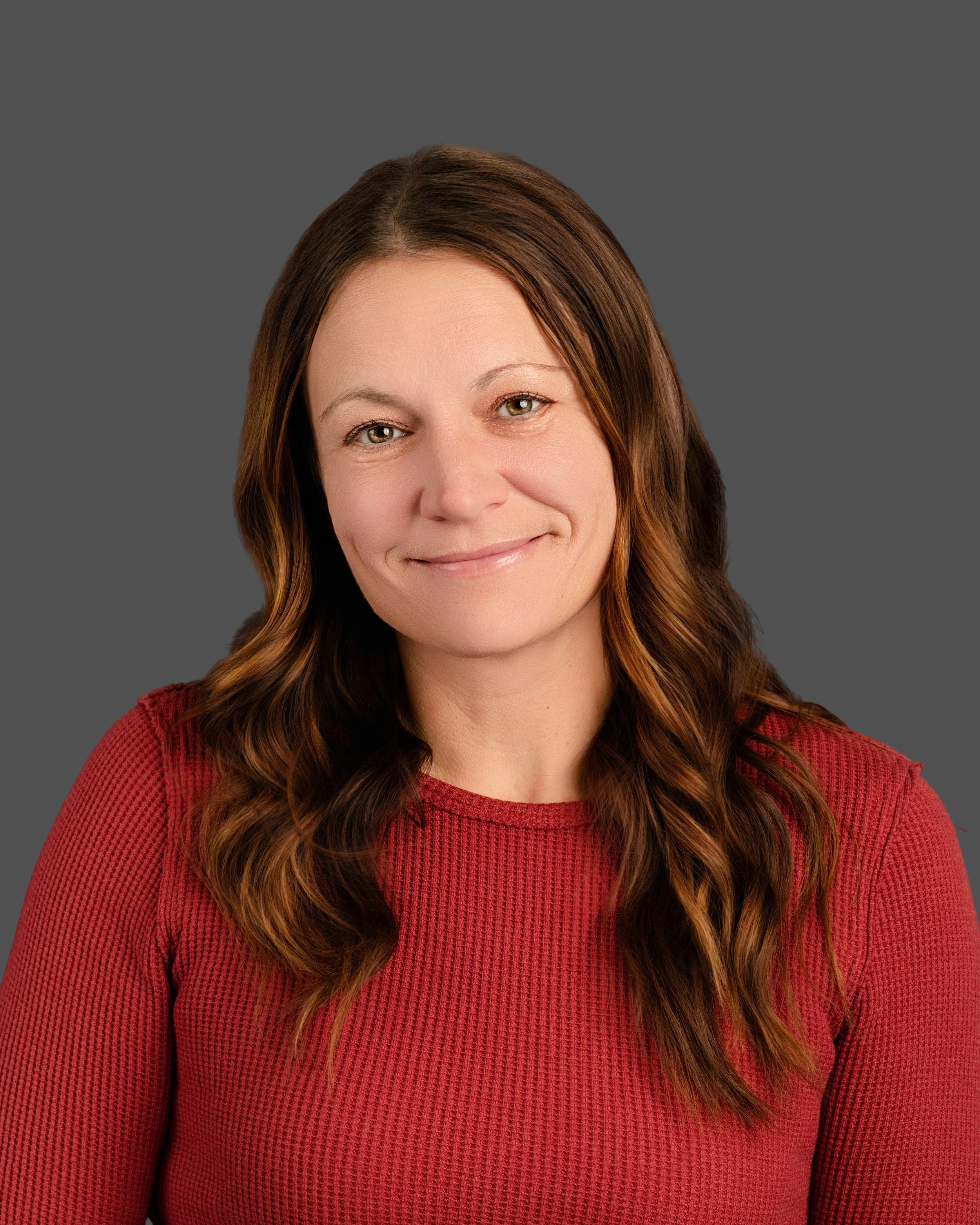 Headshot of a woman with long, wavy brown hair smiling at the camera, wearing a red textured top against a gray background.