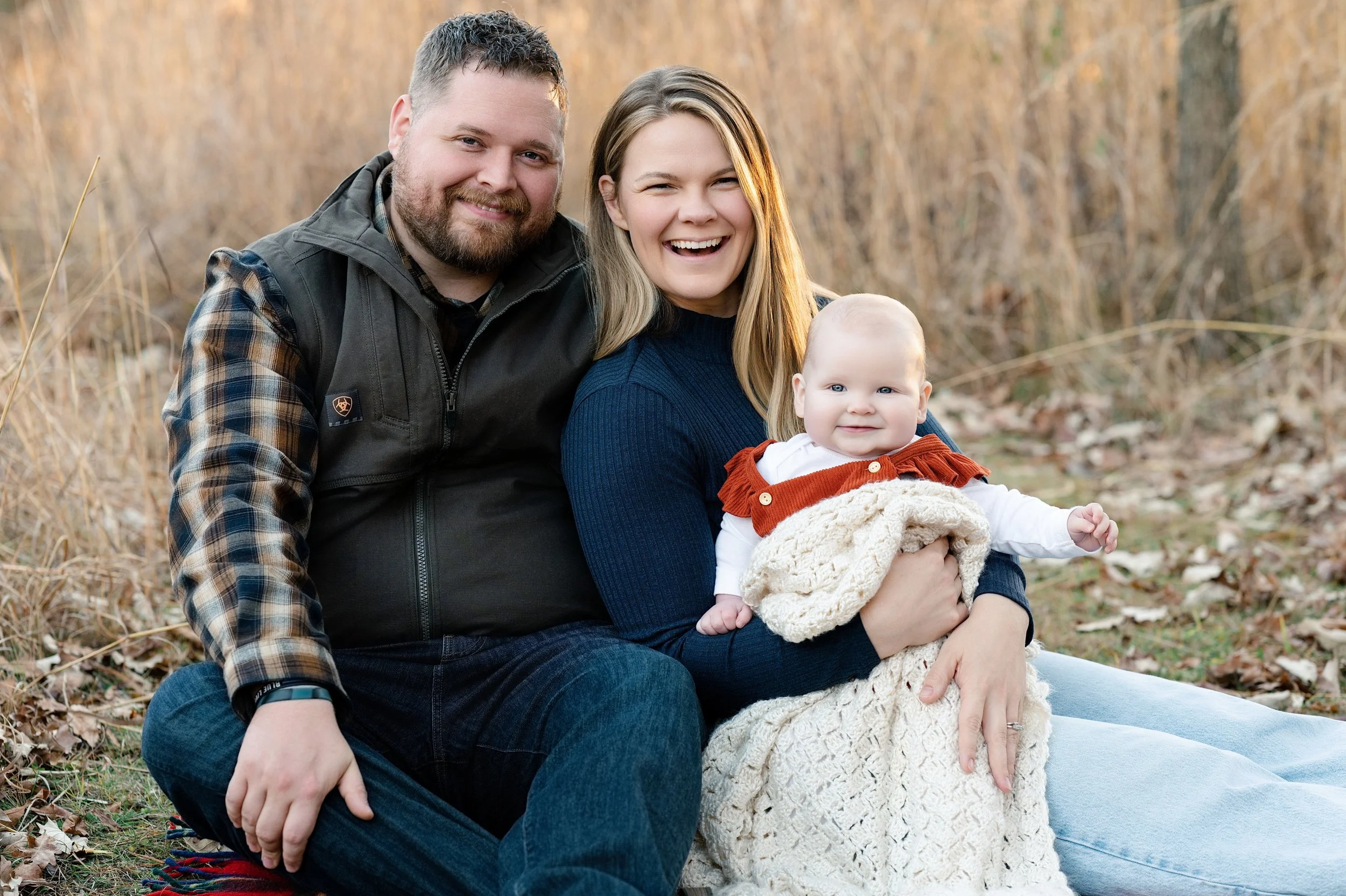 A happy family sitting outdoors in fall, with a man, woman, and a baby girl, all smiling at the camera.