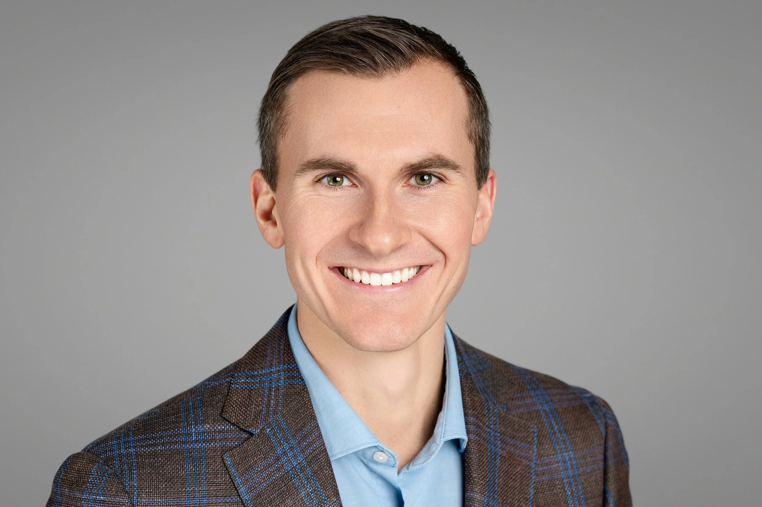 Headshot of a smiling young man with brown hair, wearing a plaid blazer and light blue shirt against a gray background.