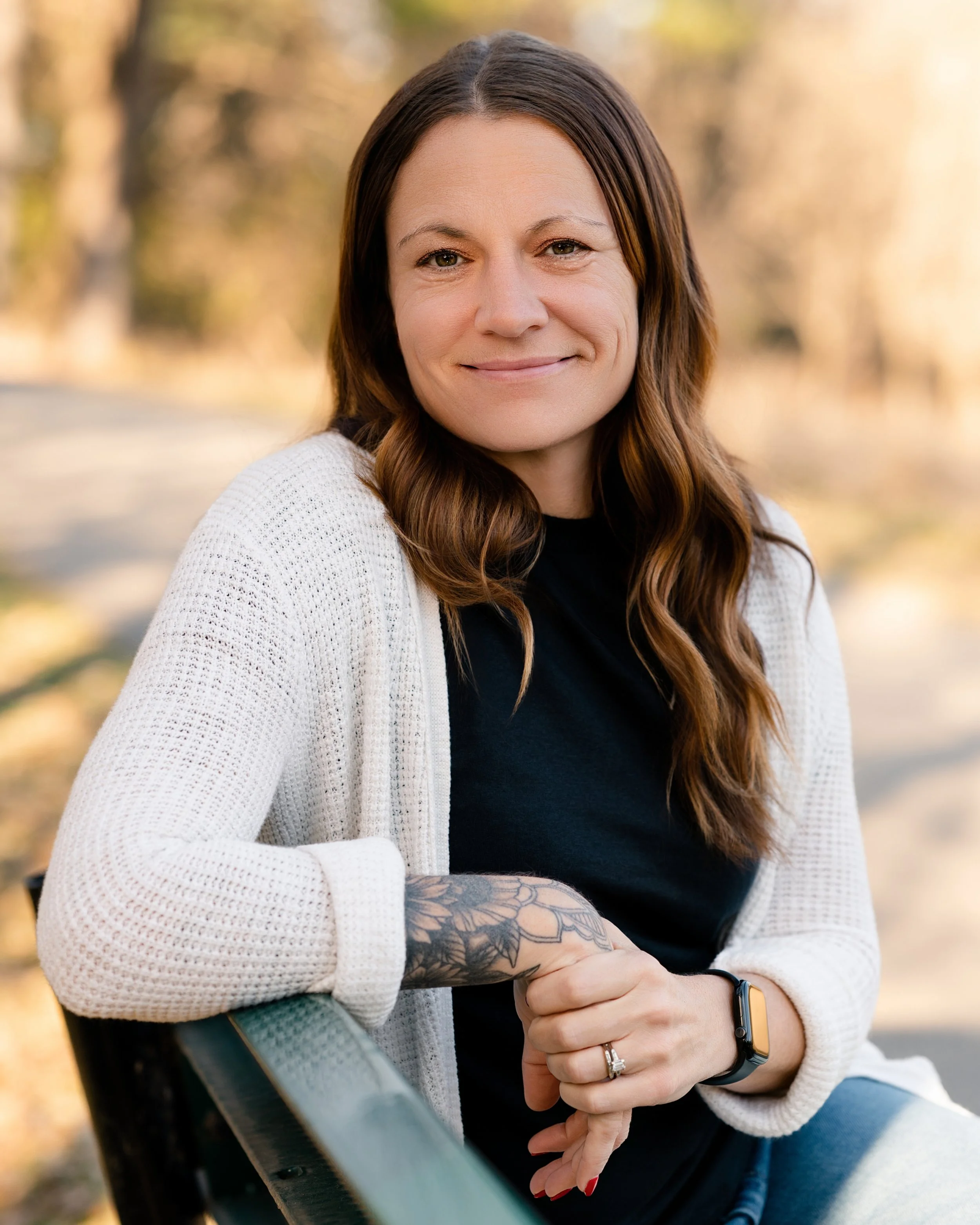 A woman with long brown wavy hair, wearing a black shirt and white knit cardigan, sitting outdoors on a bench in a park with blurred trees in the background.