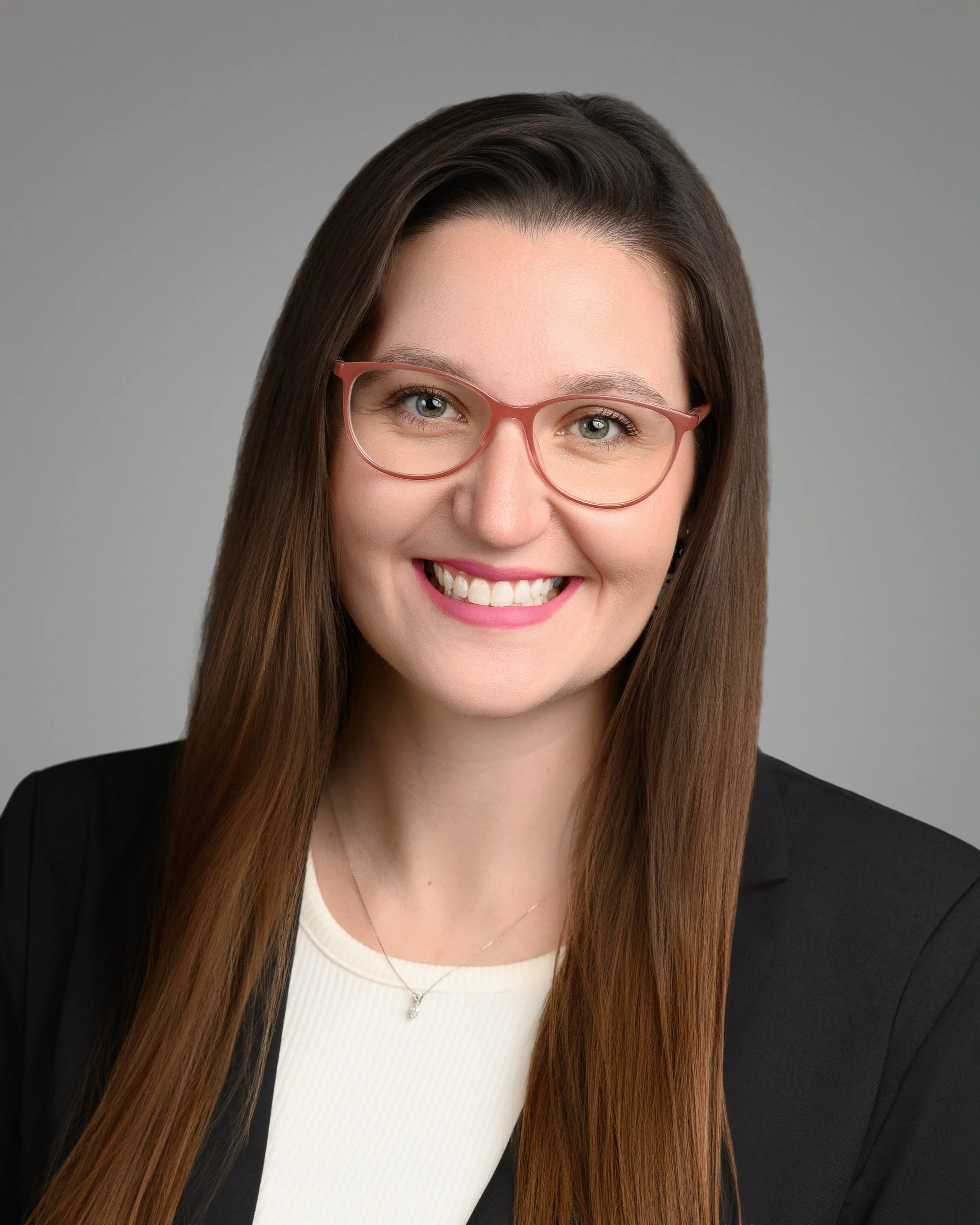 A woman with long brown hair, pink-framed glasses, and a bright smile, wearing a black blazer over a white top, against a gray background.