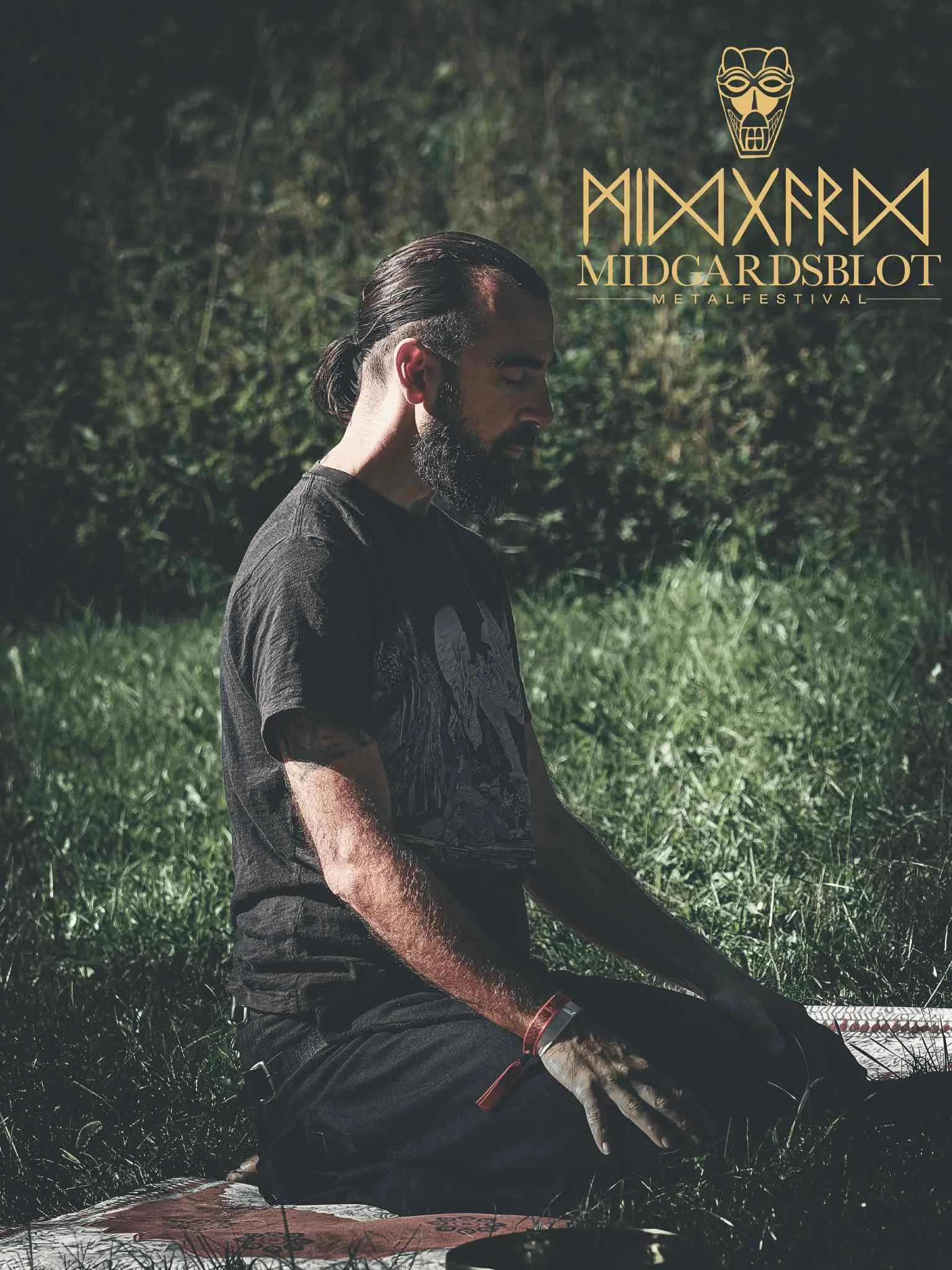 Un homme avec une barbe et des cheveux longs attachés, assis en position de méditation sur une couverture dans un espace extérieur verdoyant.