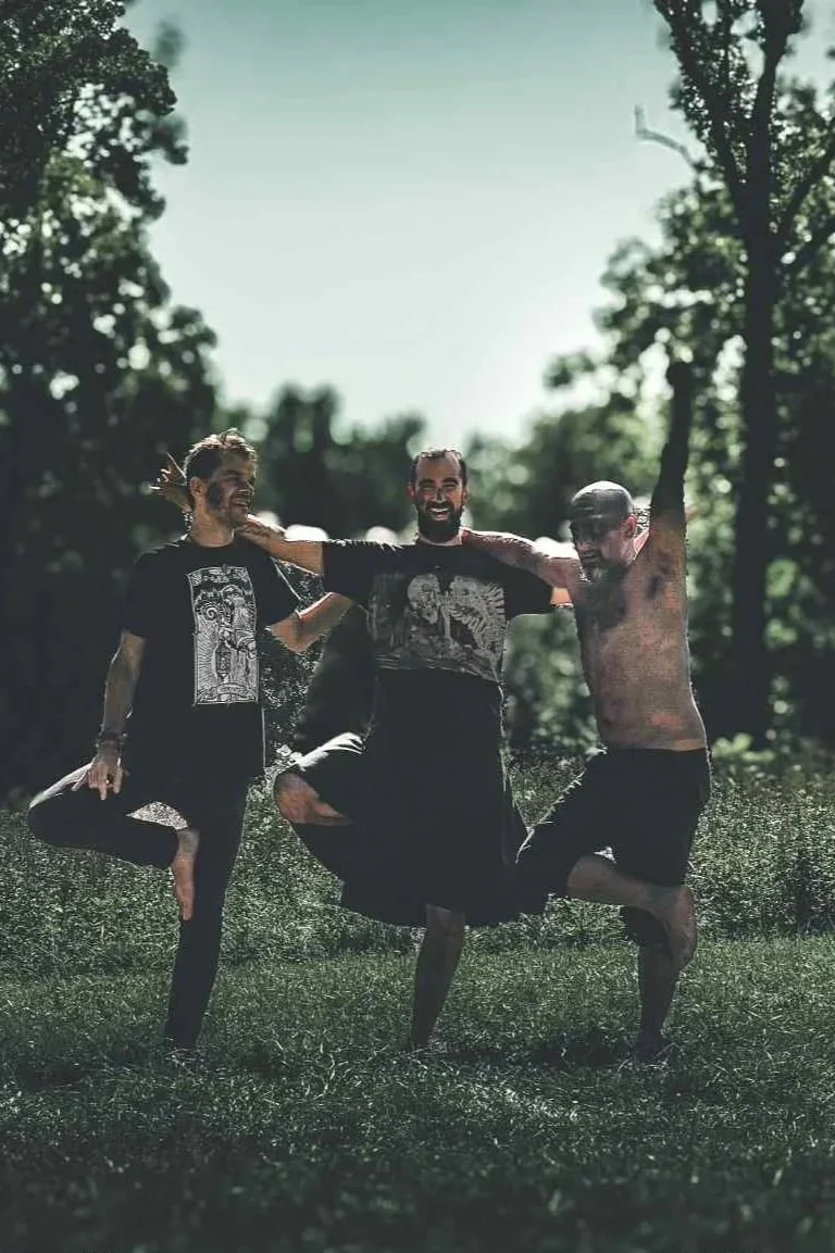 Trois hommes souriants dans un parc, s'amusant en faisant de la balançoire humaine, avec des arbres en arrière-plan.