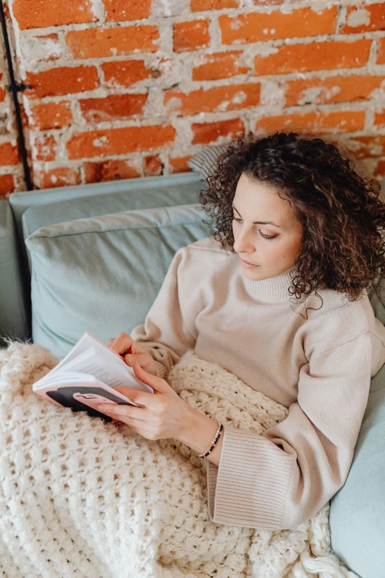 Woman with dark curly hair, laying under a blanket on the couch. She is readding a book.