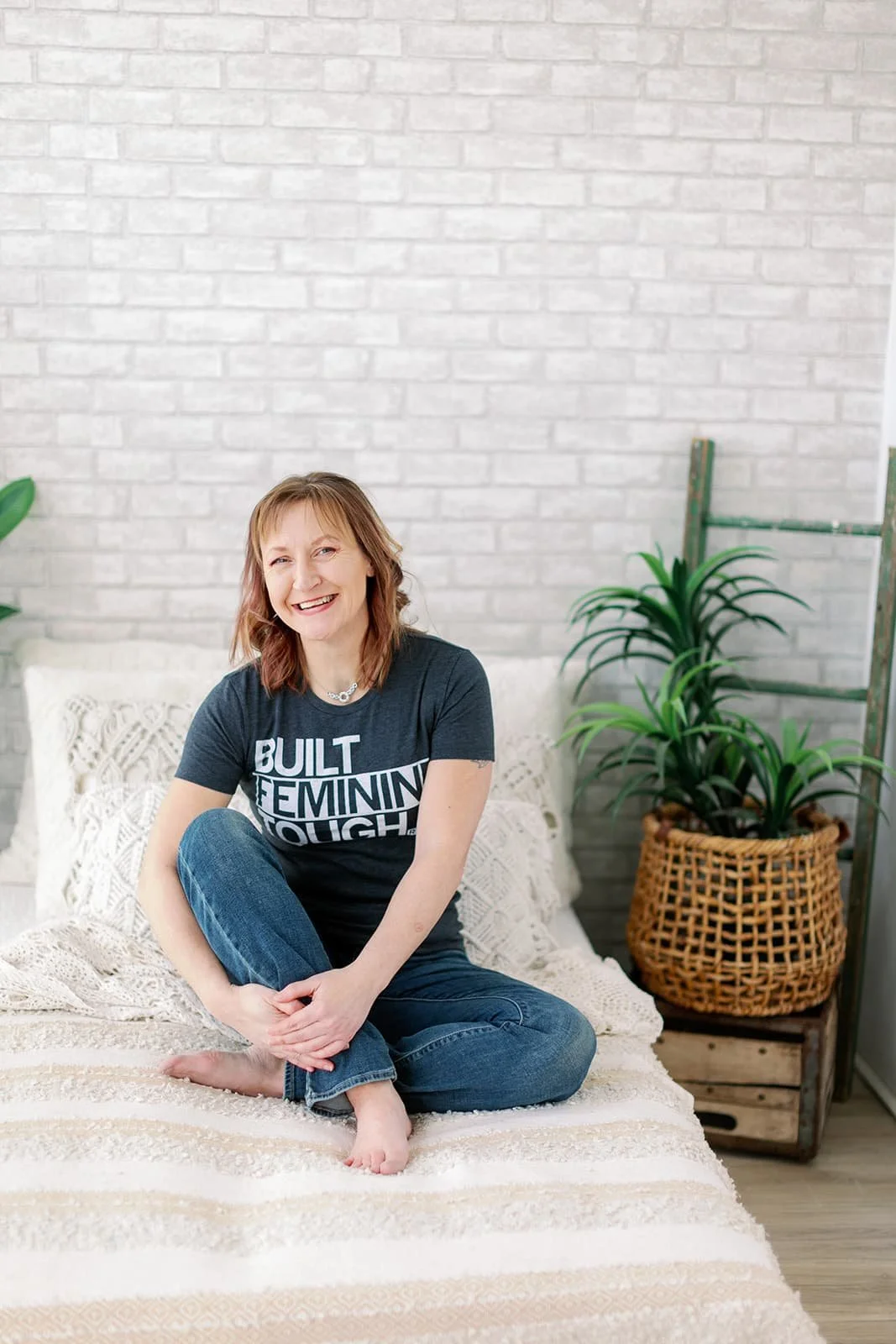 Jacki Hayes, a white woman, is sitting on a bed, with her legs crossed, one knee up, She is smiling at the camera, wearing jeans and a t-shirt.