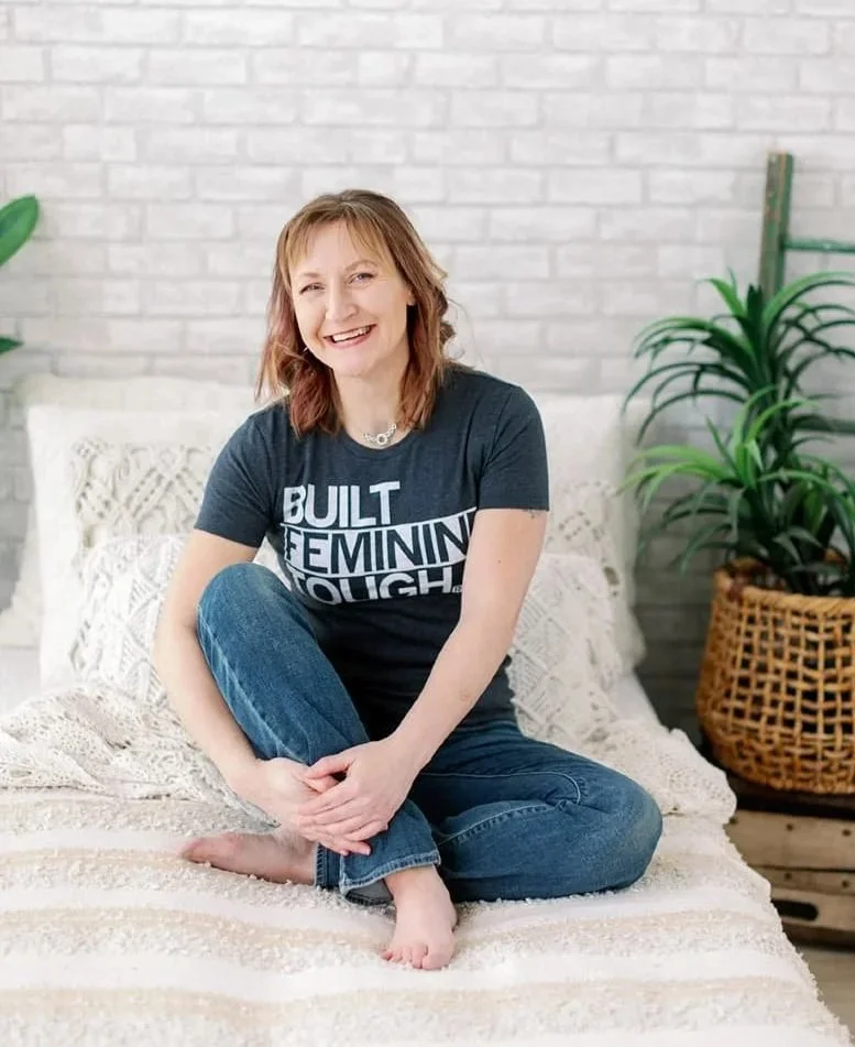 System strategist, Jacki Hayes is sitting on a bed, with her knees pulled up. She is a white woman, with brown hair, and is wearing a t-shirt and jeans.
