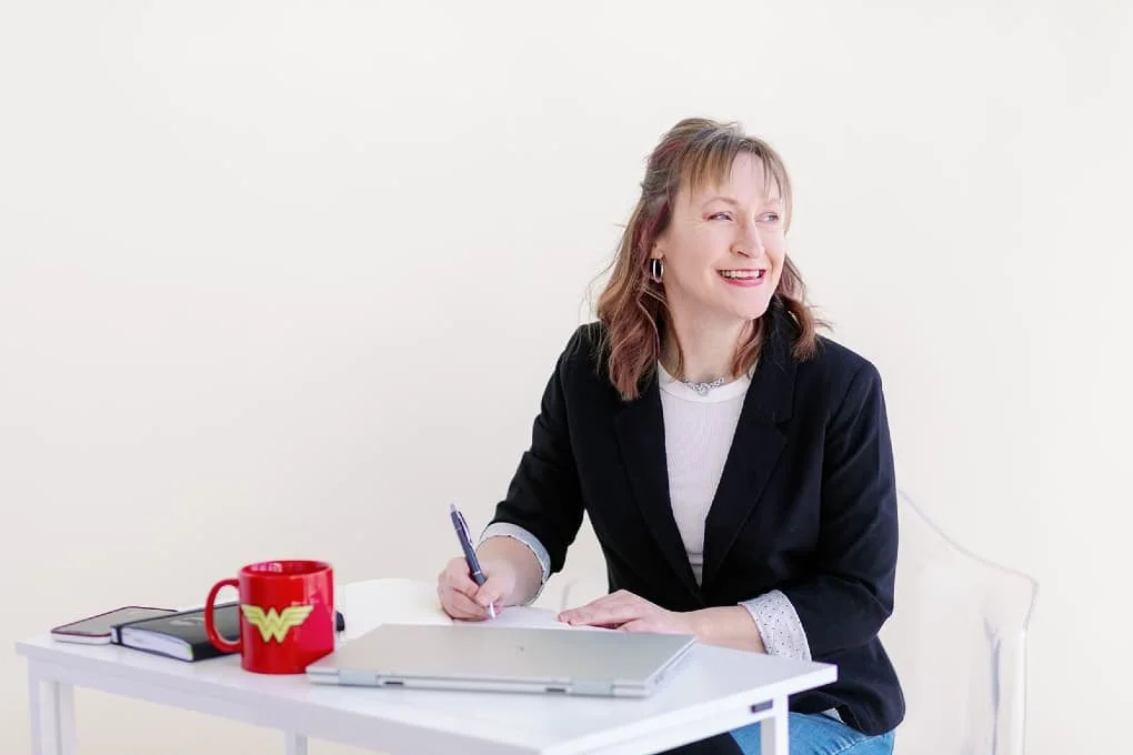 Jacki Hayes, system strategist, is sitting at a white desk writing in a notebook. There is a laptop and a red Wonder Woman mug on the desk.