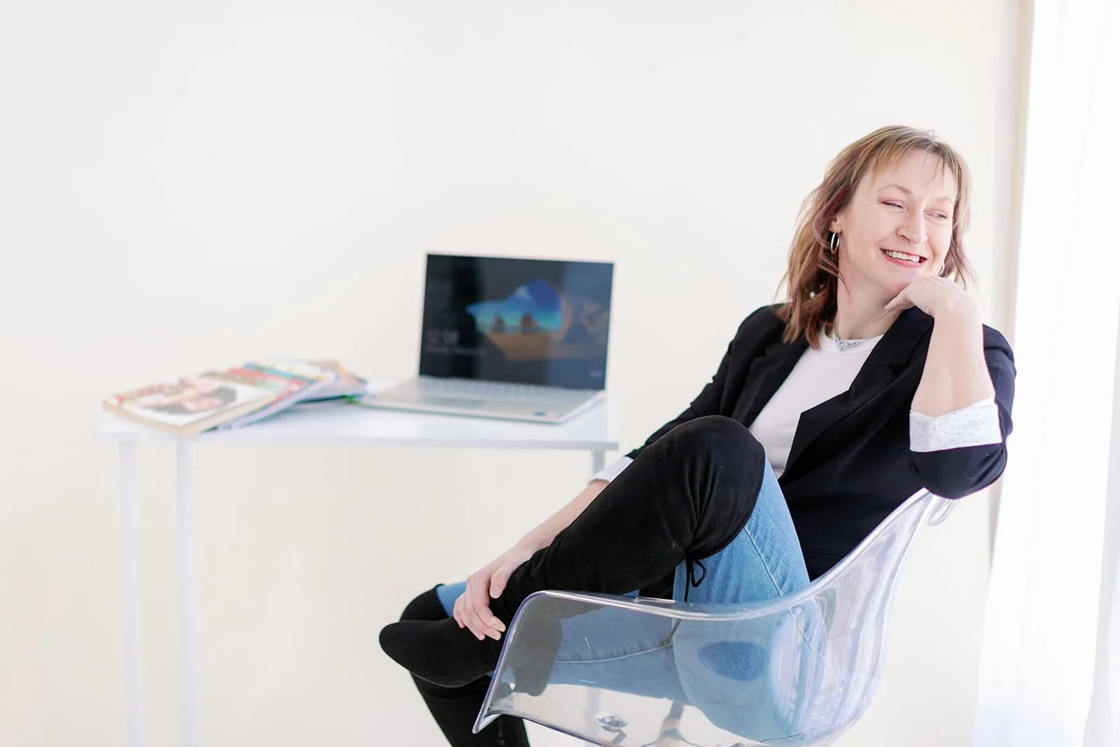 Jacki Hayes, sitting in a clear chair with a desk behind her. She is wearing a black blazer and jeans while smiling over her left shoulder.