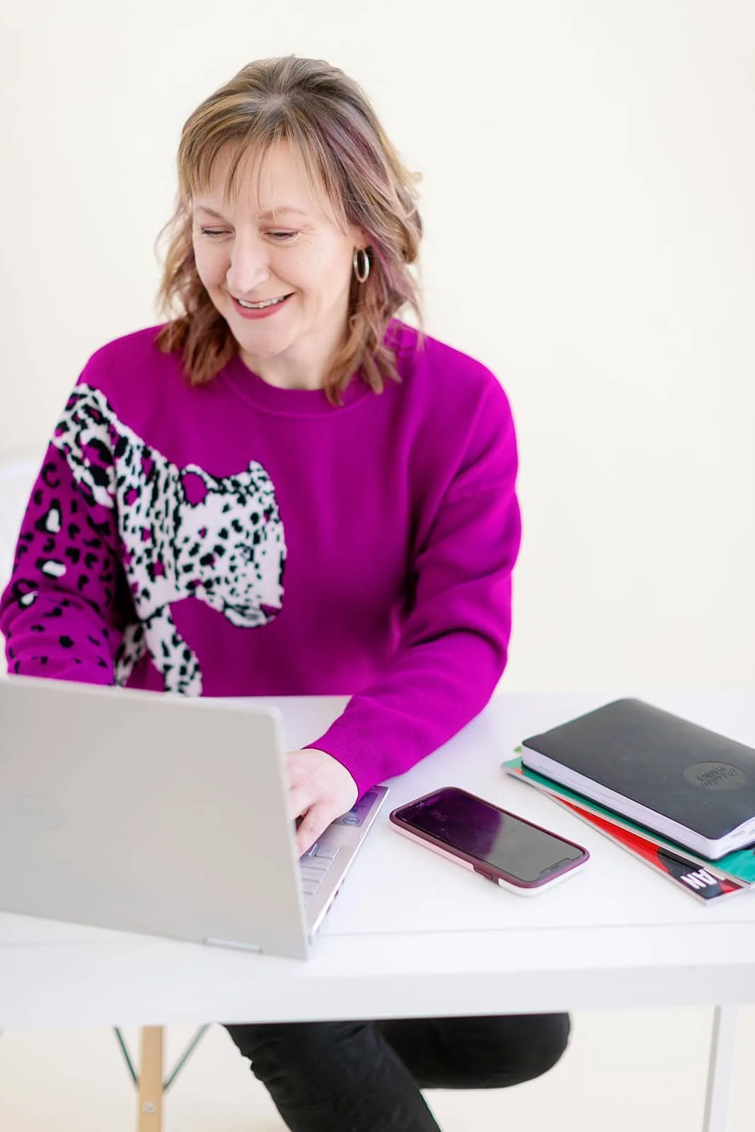 Jacki focused and smiling while typing on her laptop at a white desk, wearing her signature fuchsia leopard sweater.