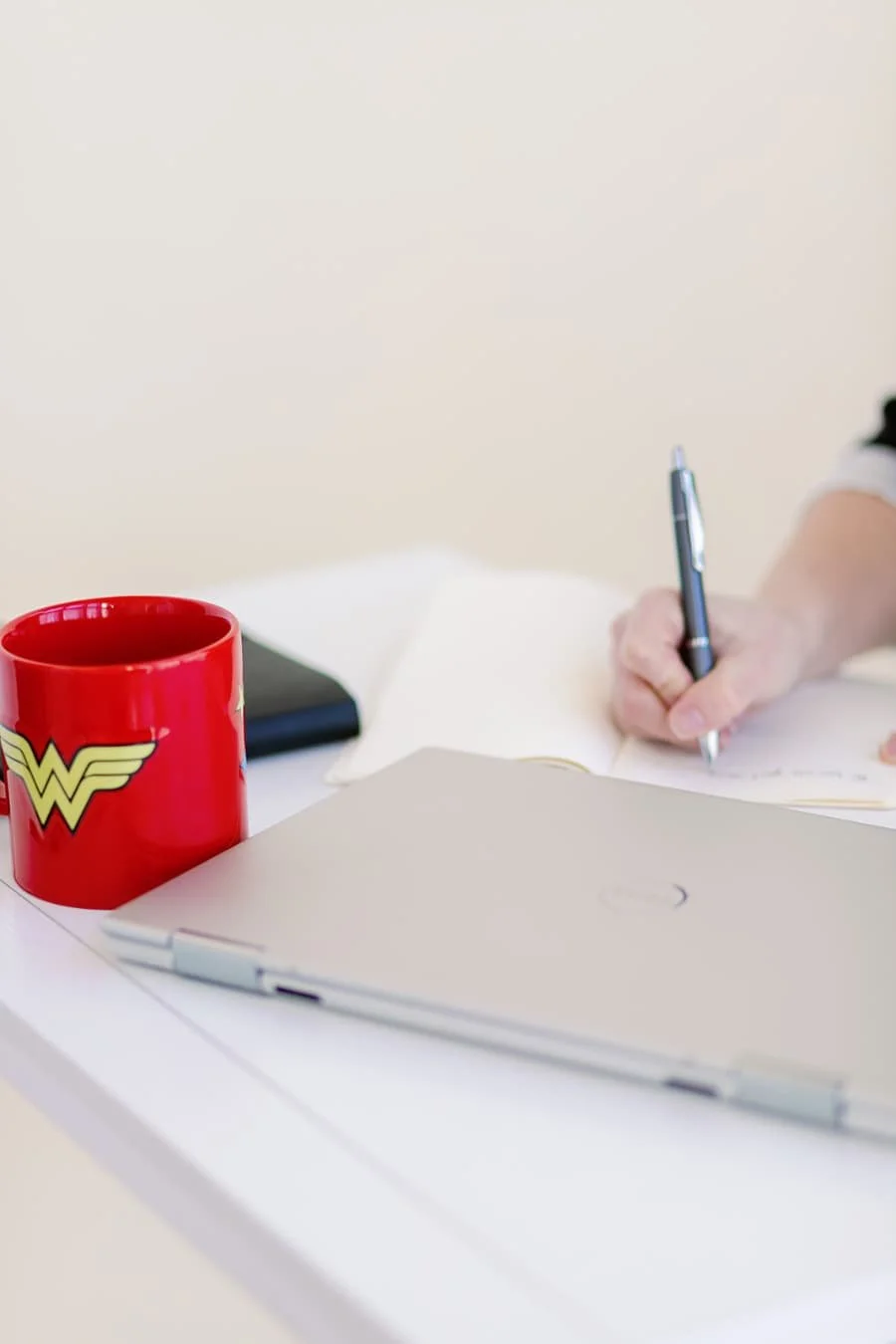 Close-up of Jacki Hayes writing in a notebook. On the desk are also a red Wonder Woman mug and closed laptop.