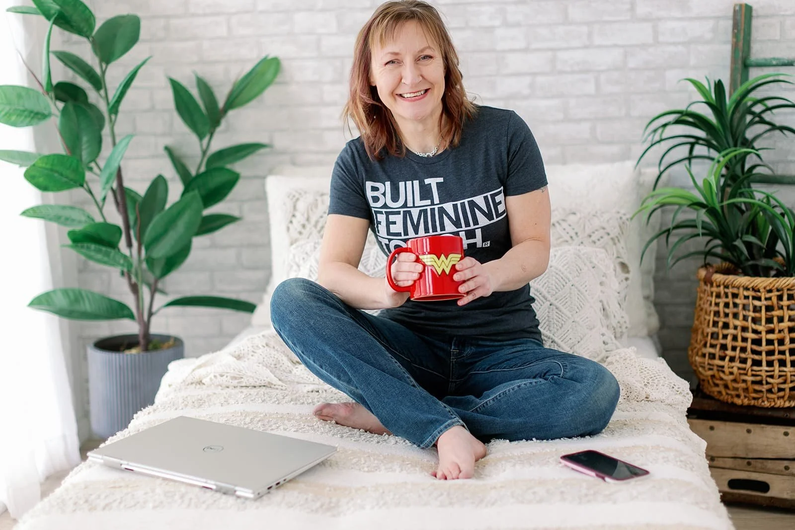 Jacki Hayes, a white woman with medium length brown hair, is sitting on a bed in jeans and a t-shirt that reads "Built Feminine Tough". She is holding a red Wonder Woman mug.