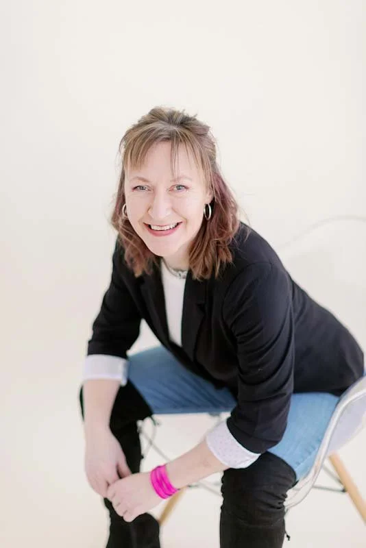 A woman with shoulder-length light brown hair, wearing a black blazer over a white shirt, and light blue jeans, sitting on a transparent chair against a plain white background. She is smiling and looking at the camera.