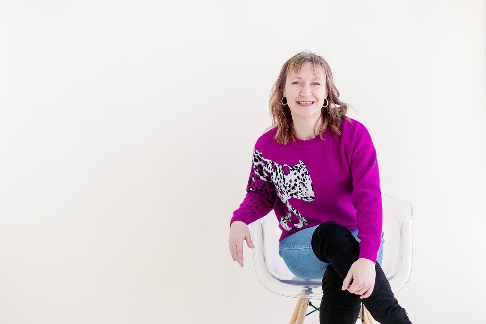 Jacki Hayes is sitting in a clear chair with her legs crossed. She is in a hot pink sweater and jeans and is smiling.