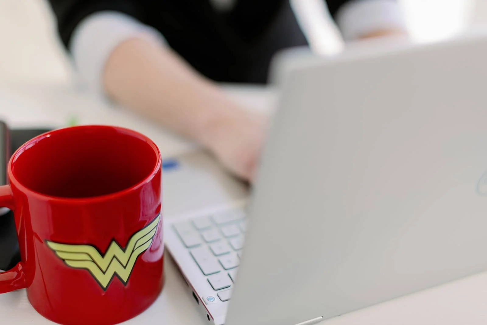 A close-up of a bright red Wonder Woman mug in the foreground with Jacki working at a silver laptop in the background. The scene captures bold focus and personal flair.