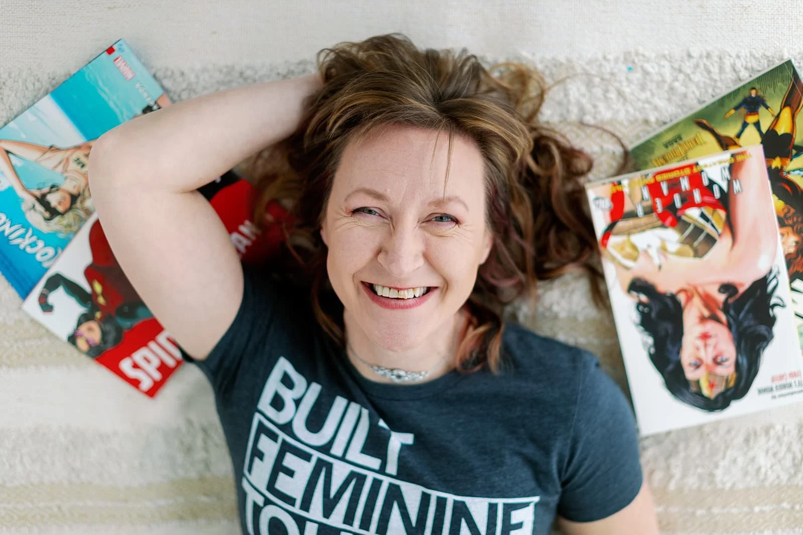 Photo of Jacki Hayes, a white woman with brown hair. She is laying face up on a bed, smiling, and surrounded by comic books. The photo is taken from above.