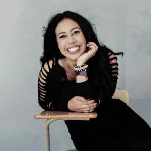 Serena Hicks smiling, resting her chin on her hand, sitting on a chair with a small table in front, wearing a black top with cut-out sleeves and jewelry, against a plain light-colored background.