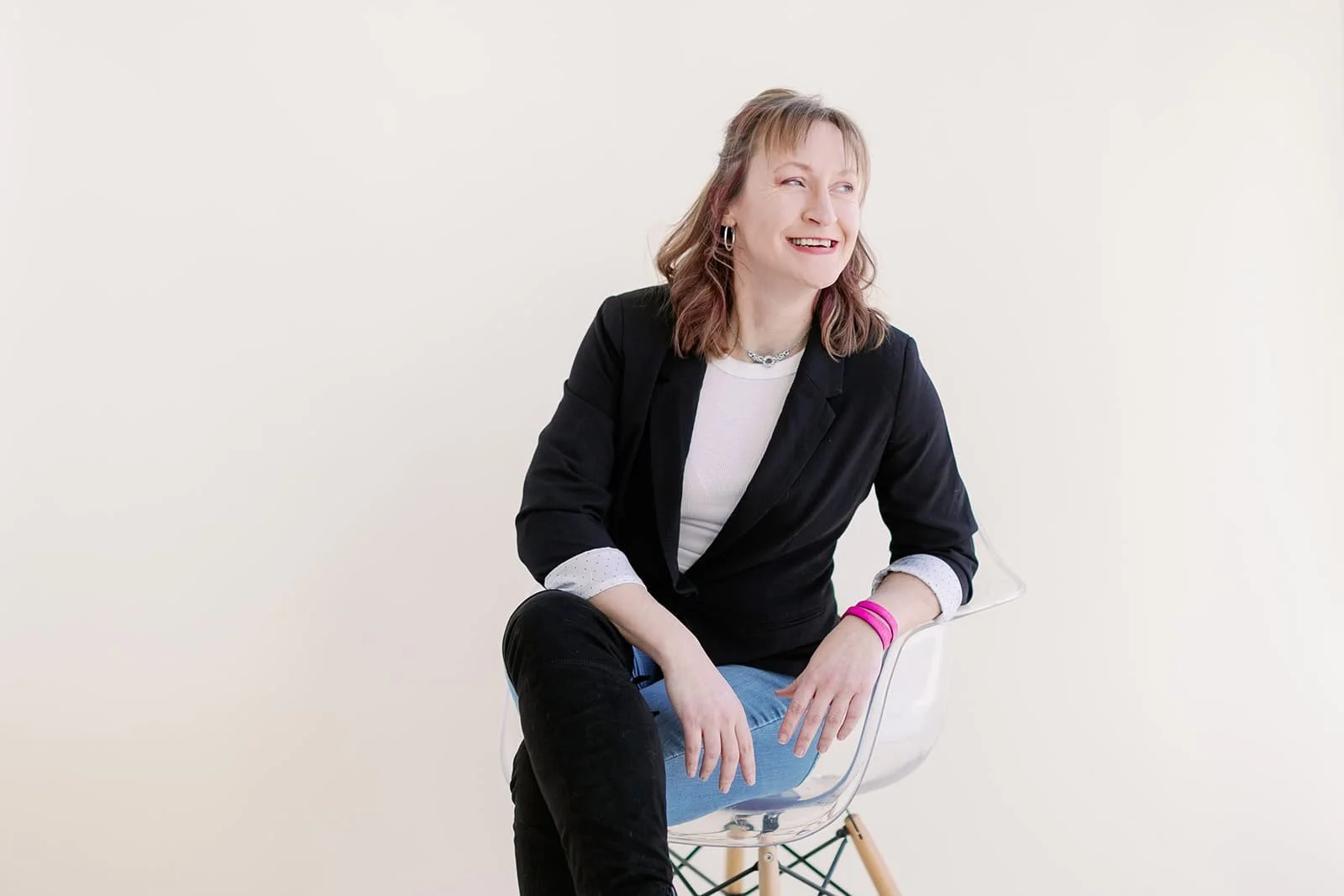 Jacki Hayes smiling and dressed in a black blazer and white shirt, sitting on a modern clear plastic chair against a plain white wall.