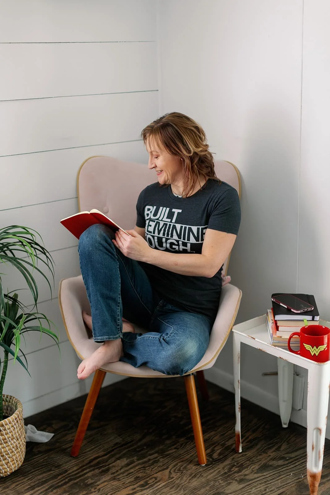 Jacki, a white woman with shoulder-length brown hair, is sitting in a modern arm chair. Her feet are up on the seat and she is writing in a journal propped on her knee.
