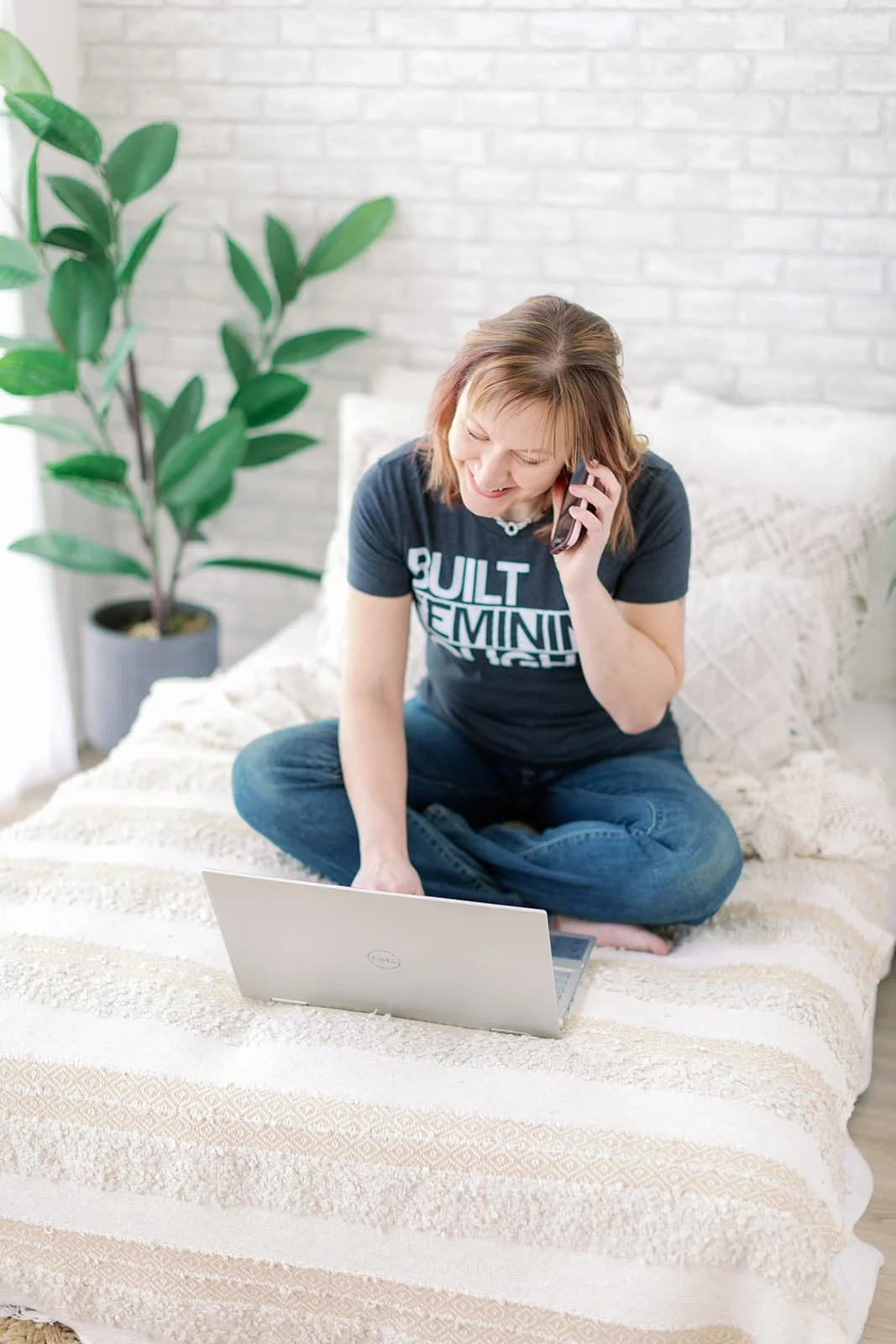 Jacki Hayes, a white woman, is sitting on bed with an open laptop. She is smiling and talking on a cell phone.