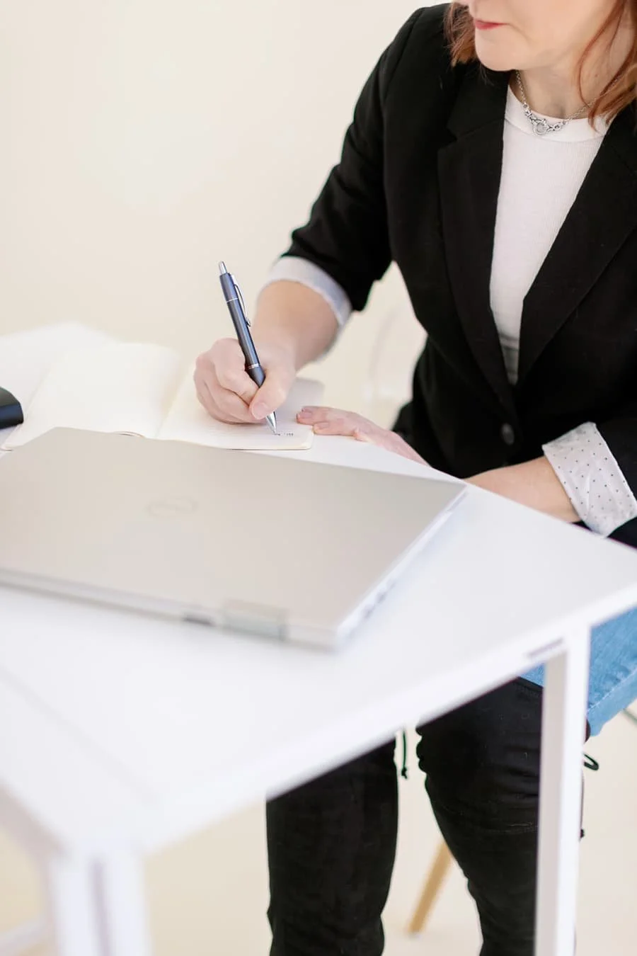 Jacki Hayes, a white woman, is sitting at a desk, writing in a notebook. In front of her is a closed silver laptop.