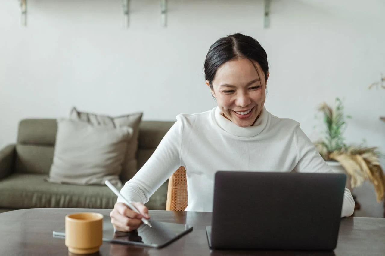 Asian woman, sitting at a table, with a couch in the background. She's looking at a computer, smiling, writing on a tablet.