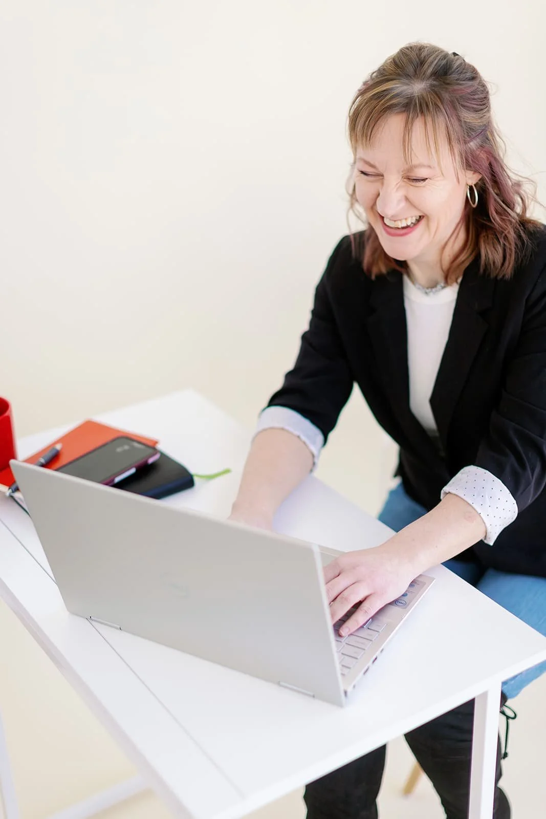 System Strategist for creative business owners, laughs while working at a white standing desk. She wears a black blazer over a white shirt, sleeves casually cuffed, and types on a silver laptop.