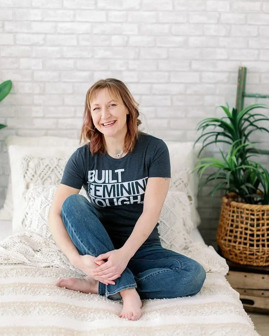 Jacki Hayes, a white woman, is sitting on a bed in jeans and a gray t-shirt.