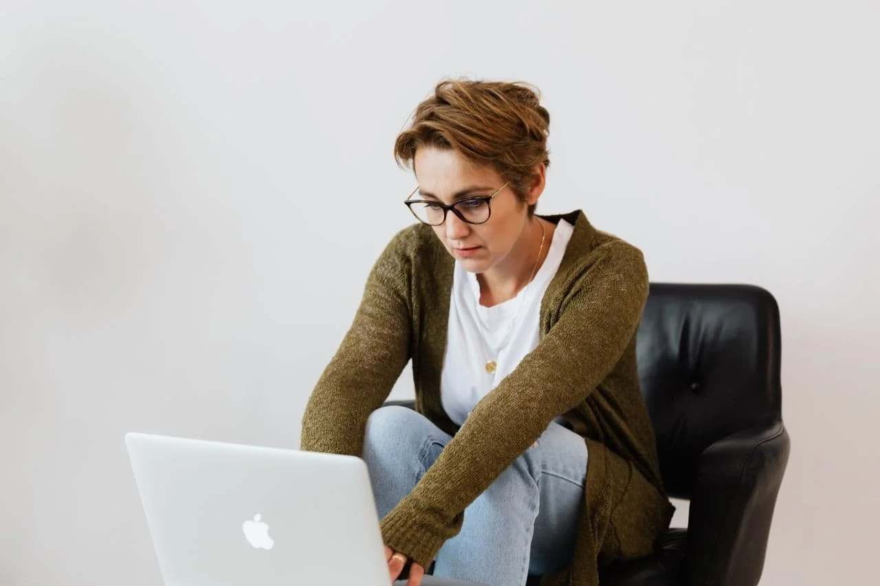 White woman, with short brown hair, sitting on an office chair, with feet on seat. She's got a laptop in front of her and she looking worried.