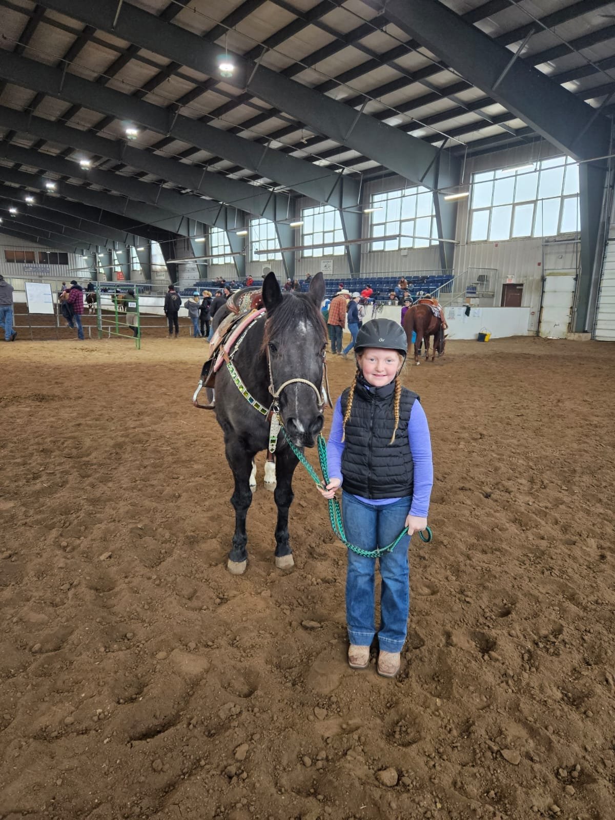 Girl with braided hair and helmet holds a black horse by a green lead rope in an indoor arena.