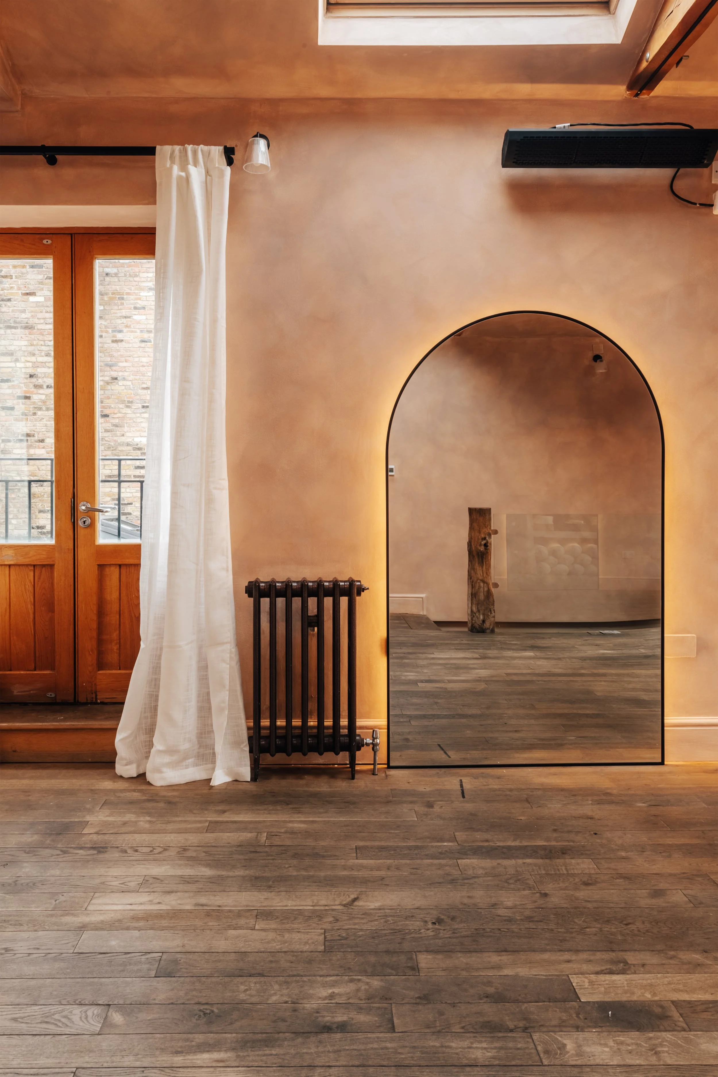 Interior view of a room with wooden flooring, a door with a glass window and white curtain, a black radiator, an archway with backlighting, and a skylight in the ceiling.