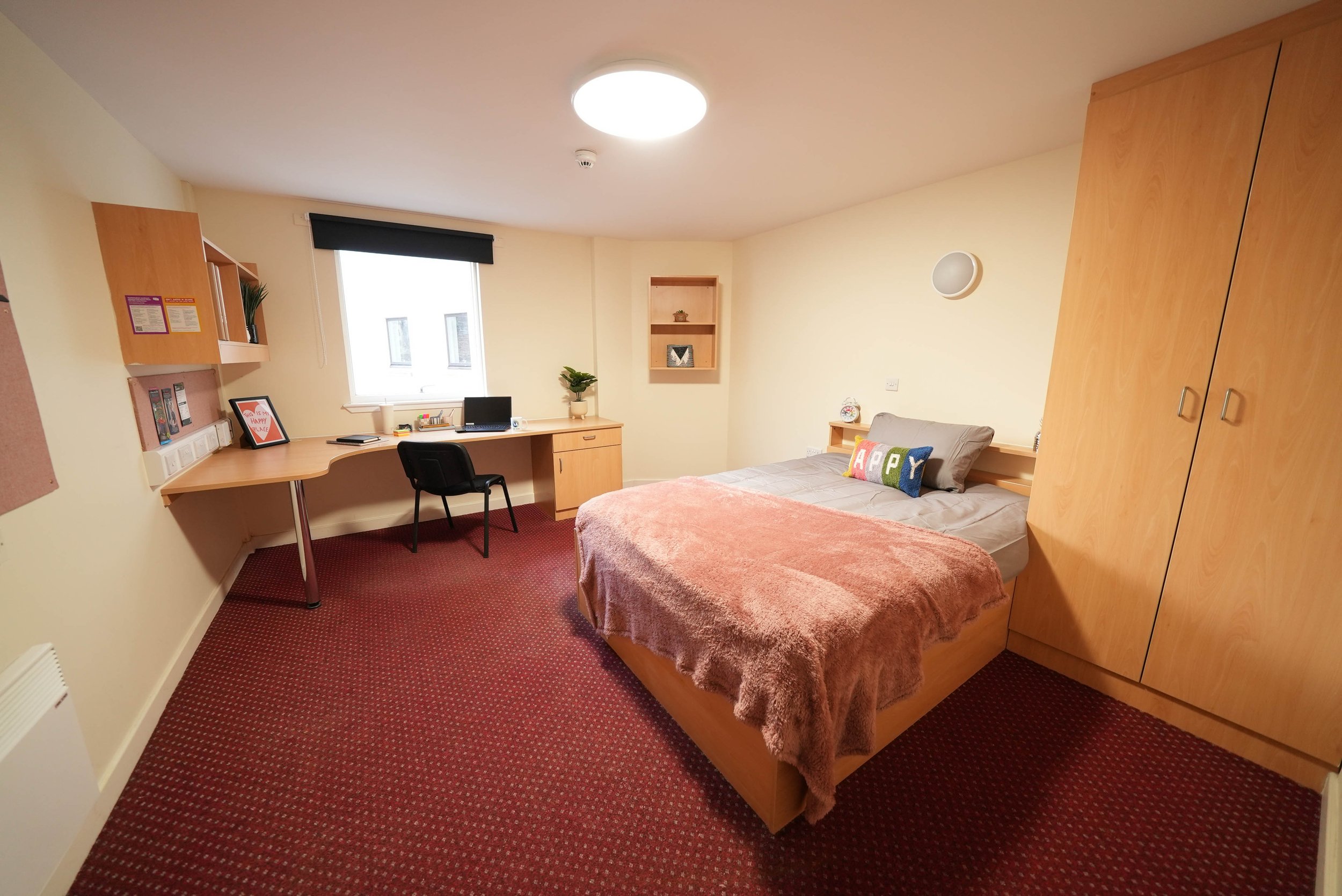 A cozy bedroom in Dundee with a bed, colorful pillow, wooden wardrobe, desk, chair, and window with blinds.