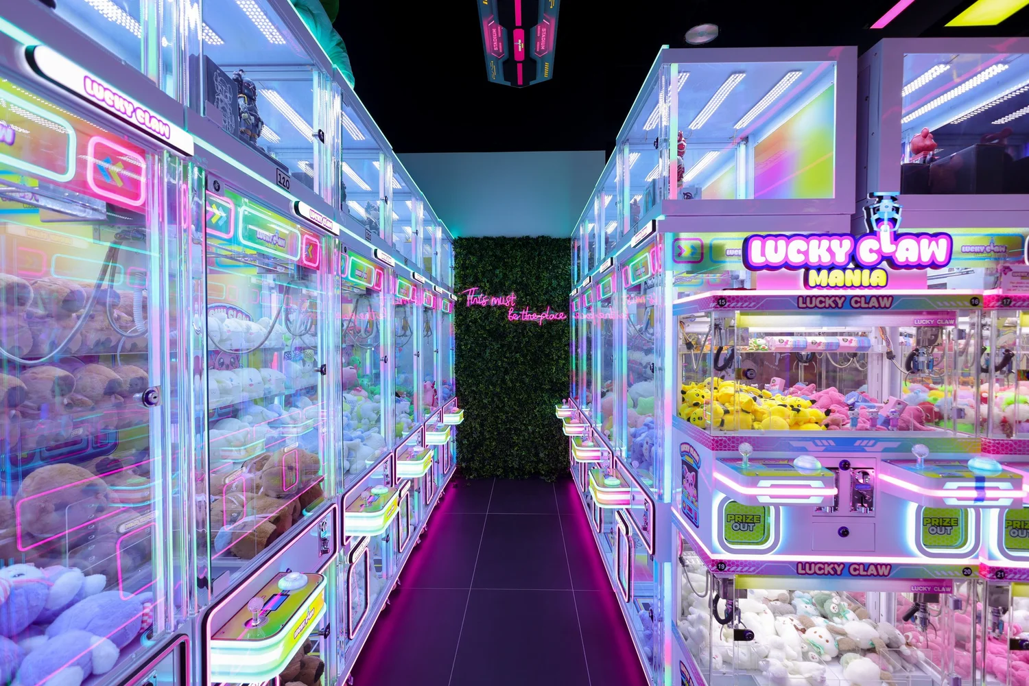 Aisle of glowing claw machines leading to a neon sign reading This must be the place