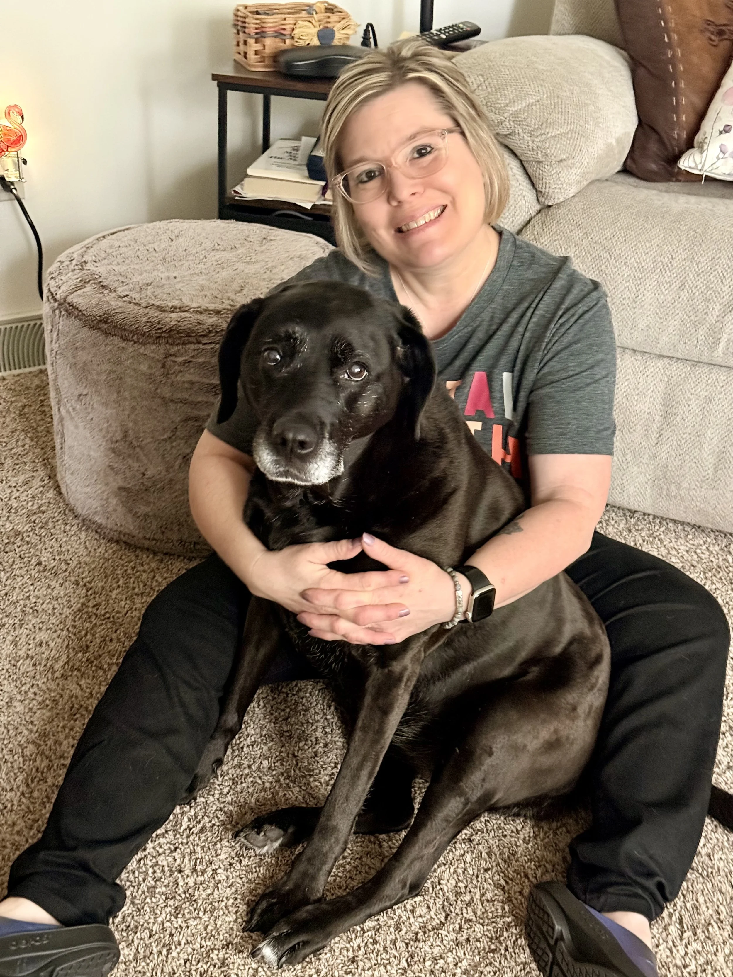 Angela sitting in front of a chair with her dog Koko, a black lab.