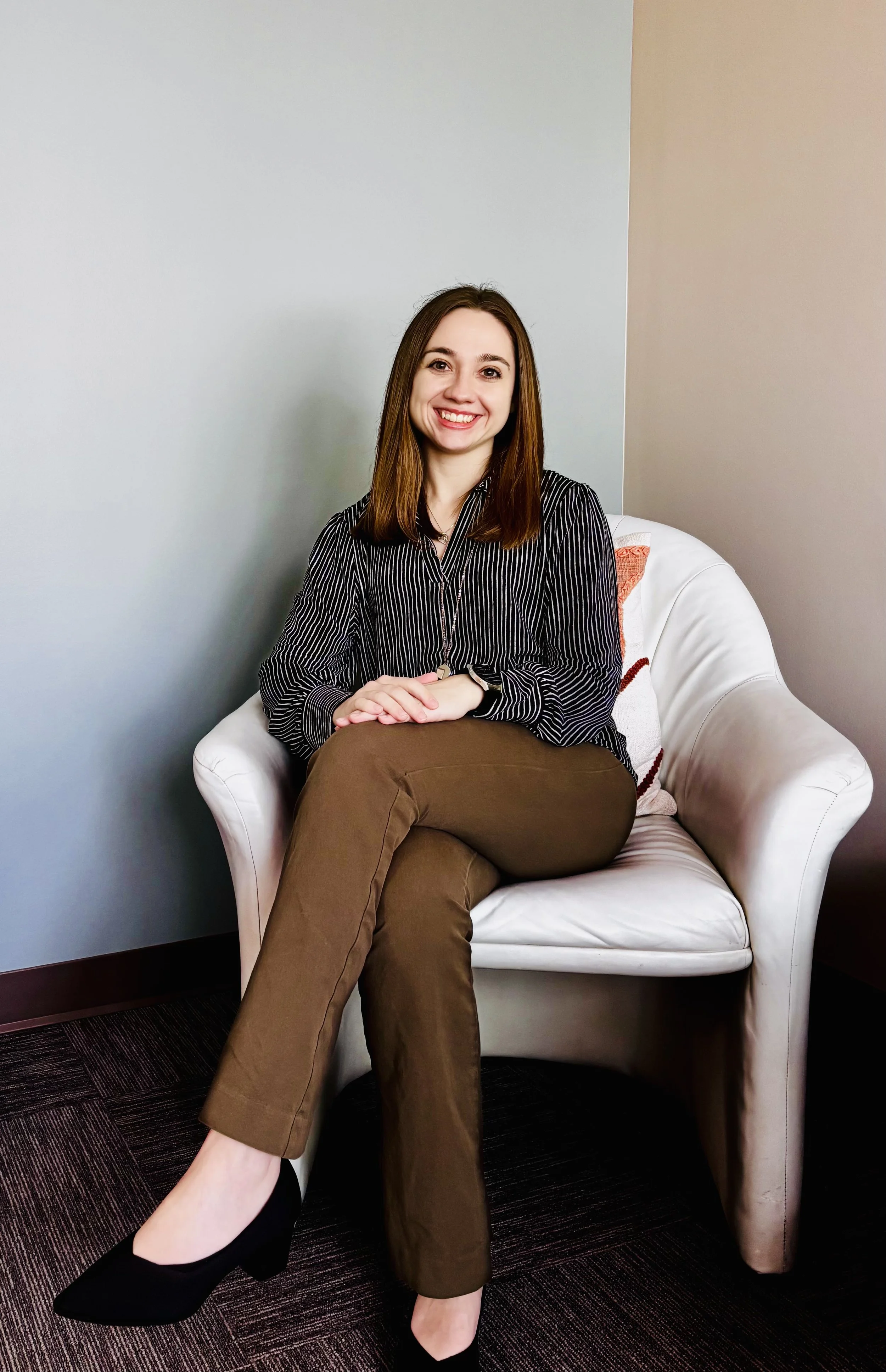 Megan Porter, smiling while sitting in a chair in her office. She has shoulder length straight-brown hair. She is wearing a black blouse with white lines and brown pants.