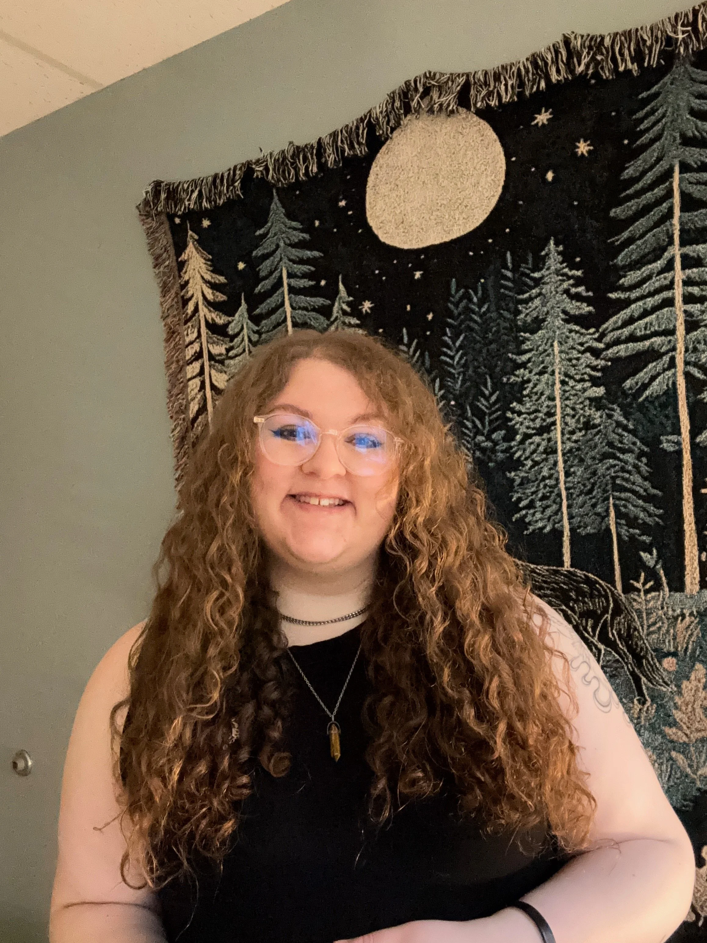 Dana Bejger has long curly light brown hair, glasses, and is wearing a sleavless black dress. She is standing against a wall hanging picture of a wolf in a forest on a moonlight night.