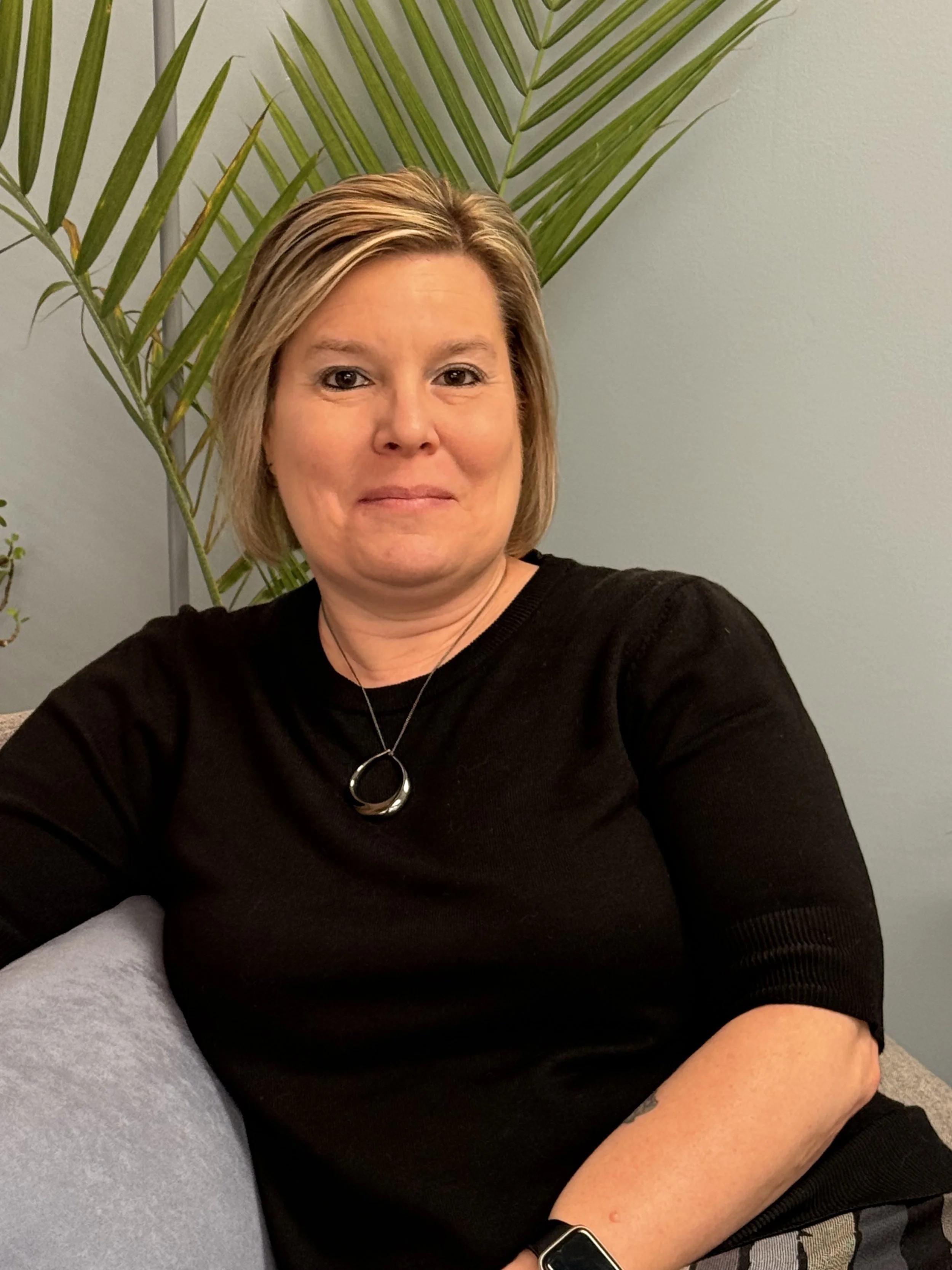 Angela Rodgers. Wearing a black sweater and siting on a grey chair in her office.