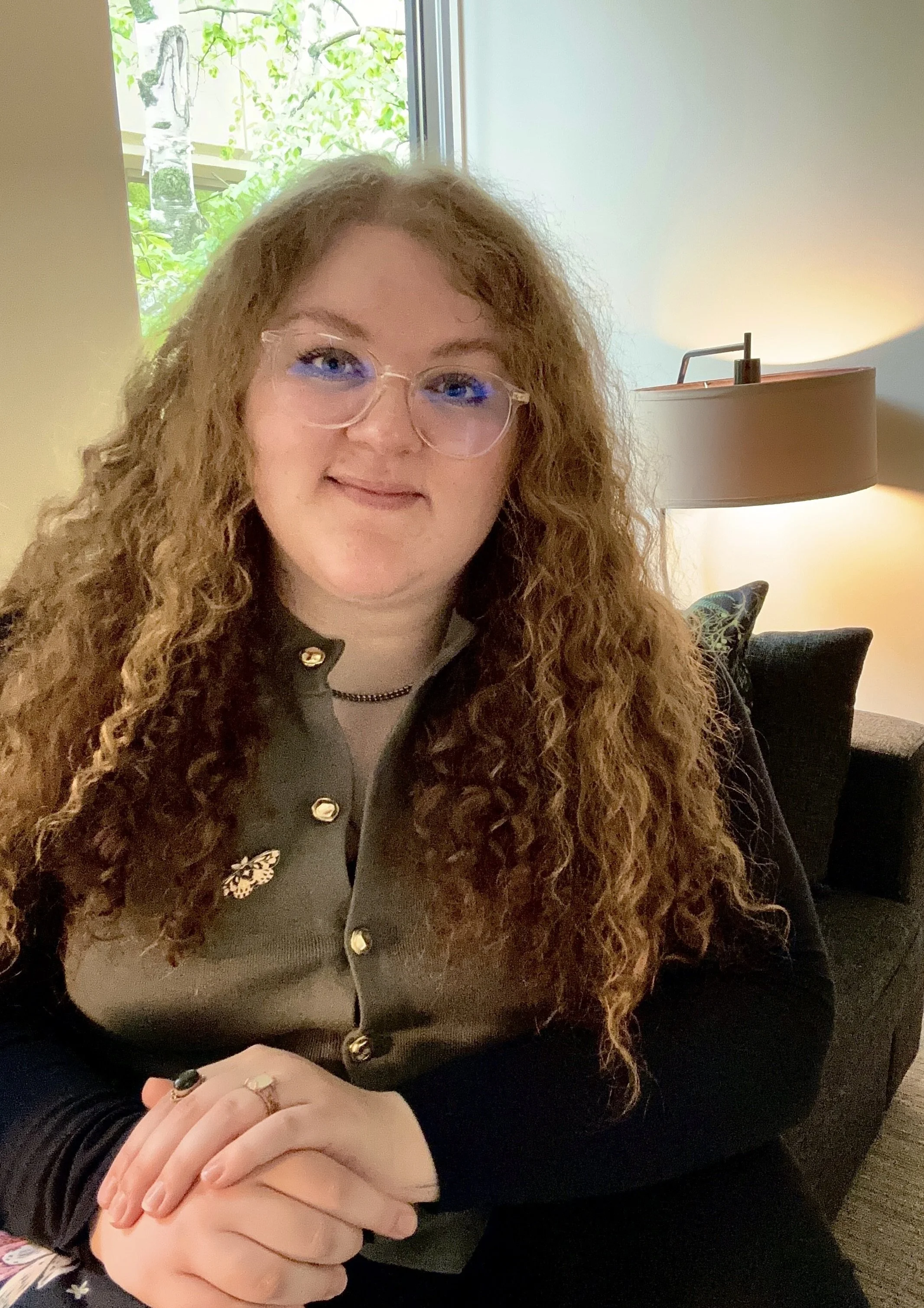 Headshot of Dana Bejger, LPC. She has long curly hair and round glasses. She is smiling at the camera with her hands folded in front of her.