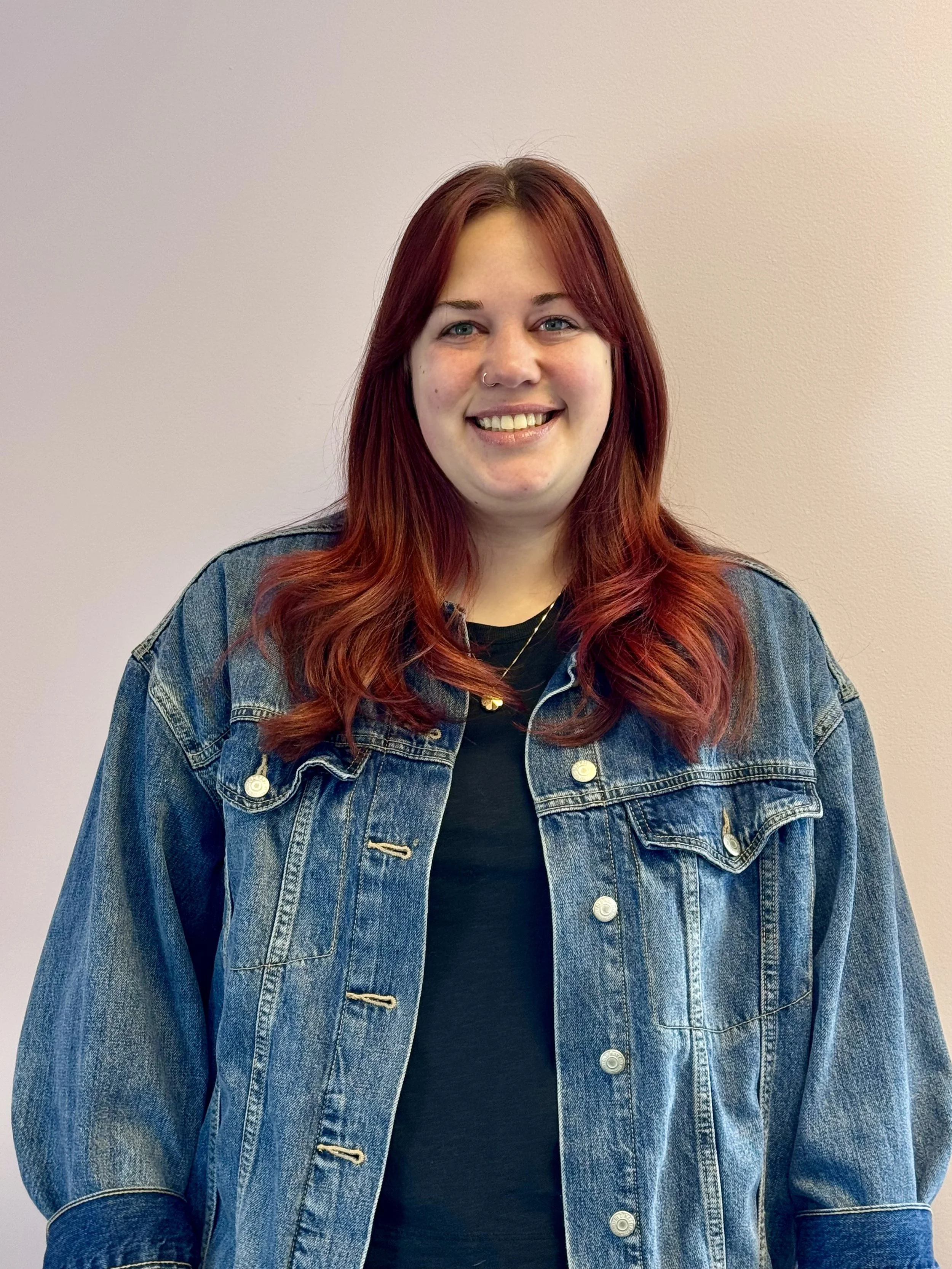 A woman in her twenties with shoulder length red hair. She is smiling and wearing a jean jacket, dark shirt and gold pendant around her neck, standing up against a light colored wall.
