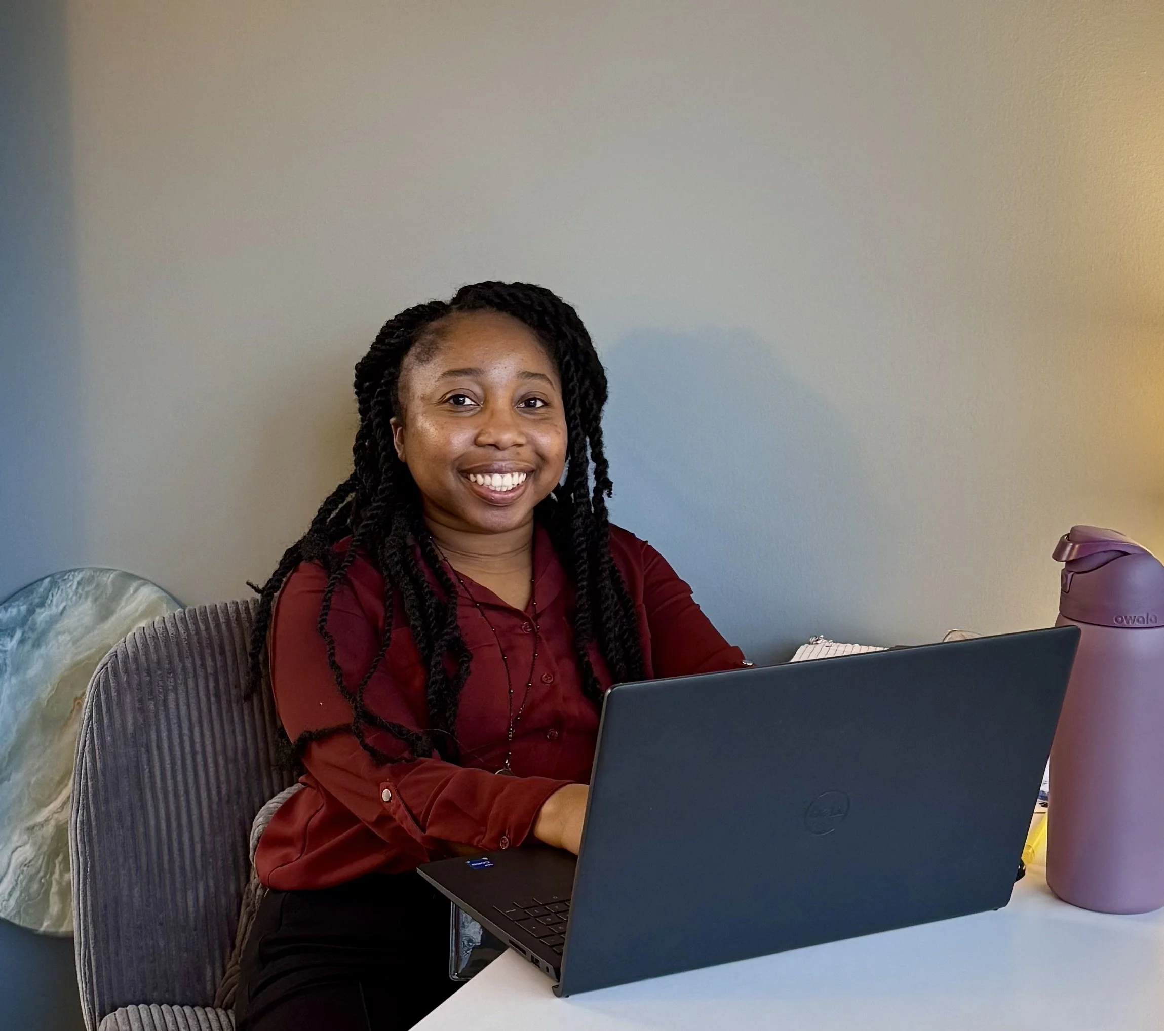 Rachel Foster sitting at her desk smiling while working on a laptop. She has long braided hair and is wearing a maroon blouse.