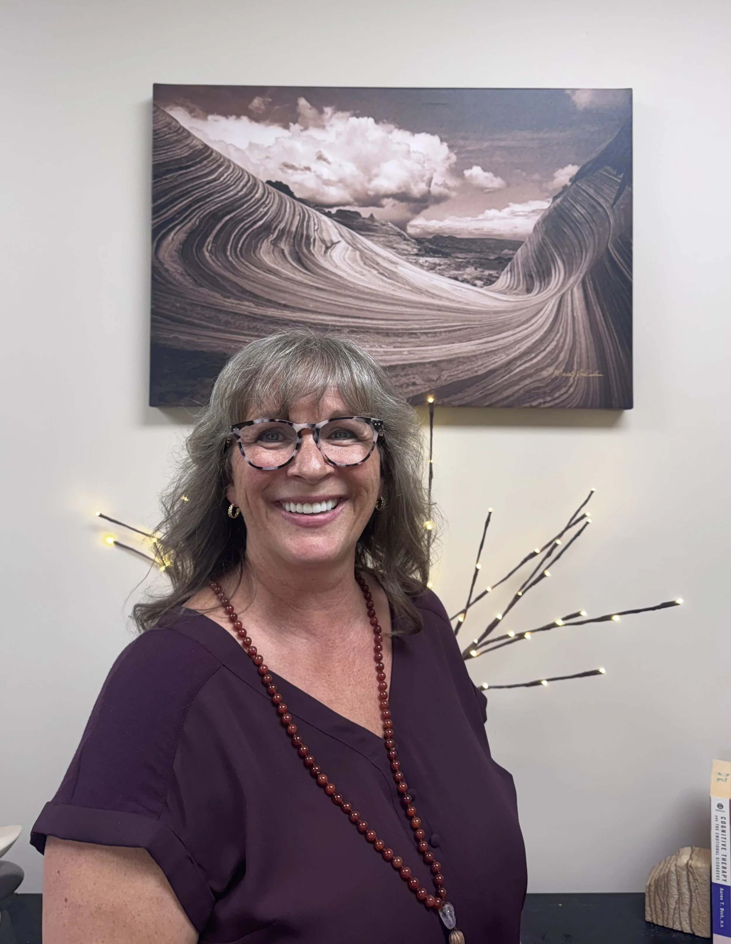 Vicki Shane wearing a purple shirt with beaded necklace. She has dark grey hair and glasses and is standing under a picture of Sandstone rock from Antelope Canyon, AZ.